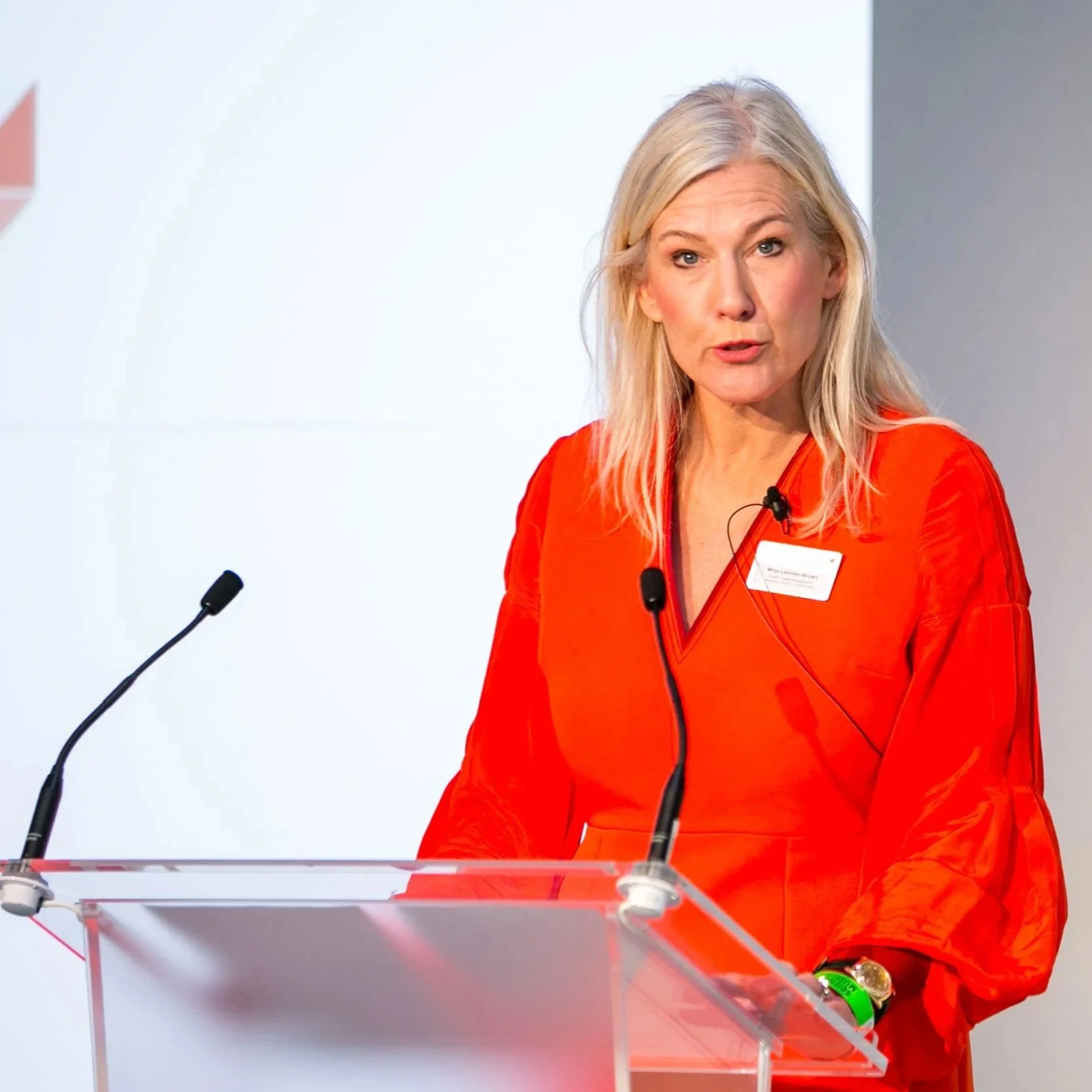 A blonde woman in a red dress speaking at a podium with microphones in front of a plain white wall.