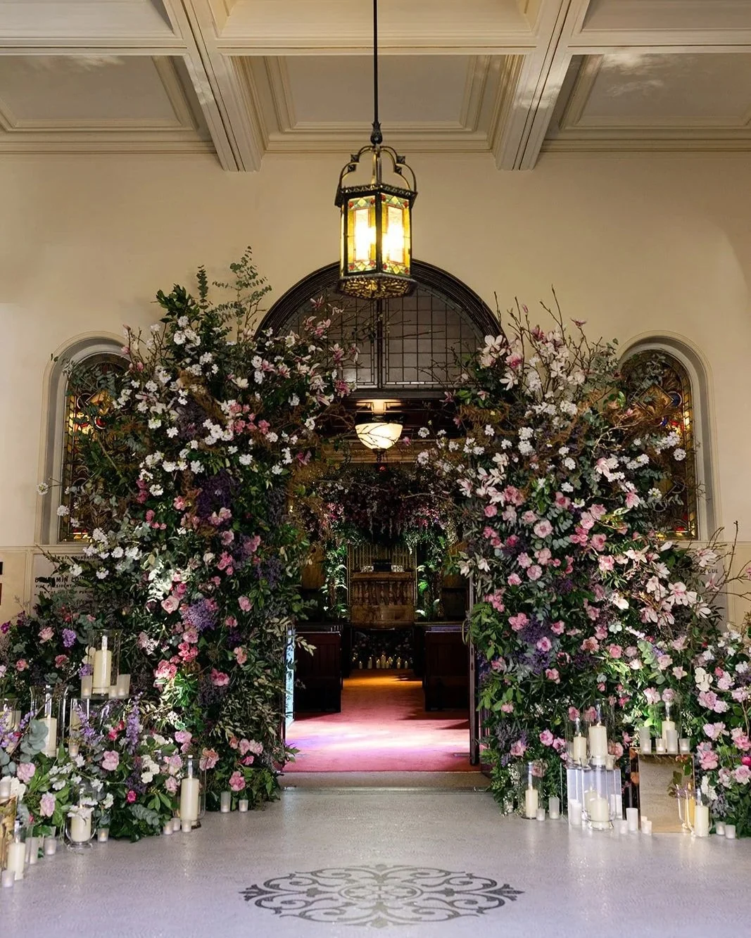 Entrance decorated with abundant pink and white flowers and candles, leading into a hall with stained glass windows and artistic lighting. Wedding at The Peninsula London