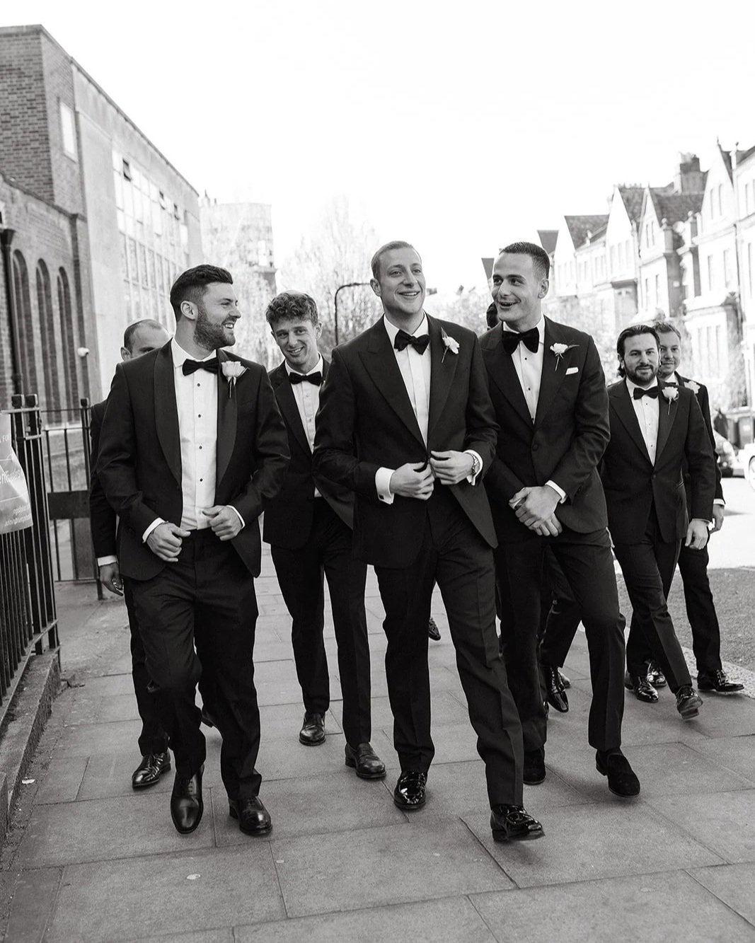 Group of men in tuxedos walking on the sidewalk, smiling, with historic buildings in the background. Walking to The Peninsula London