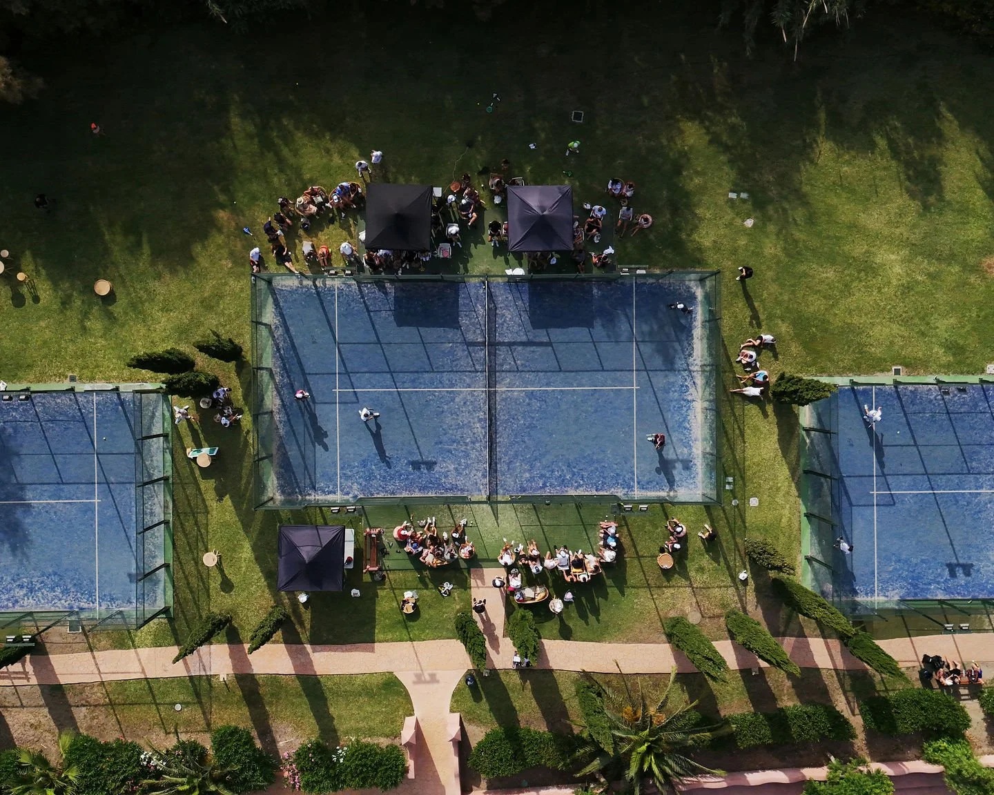 Aerial view of a sports facility with multiple padel courts and a gathering of people on the grass outside, some under black tents, during daytime at Anantara Villa Padierna Resort in Marbella