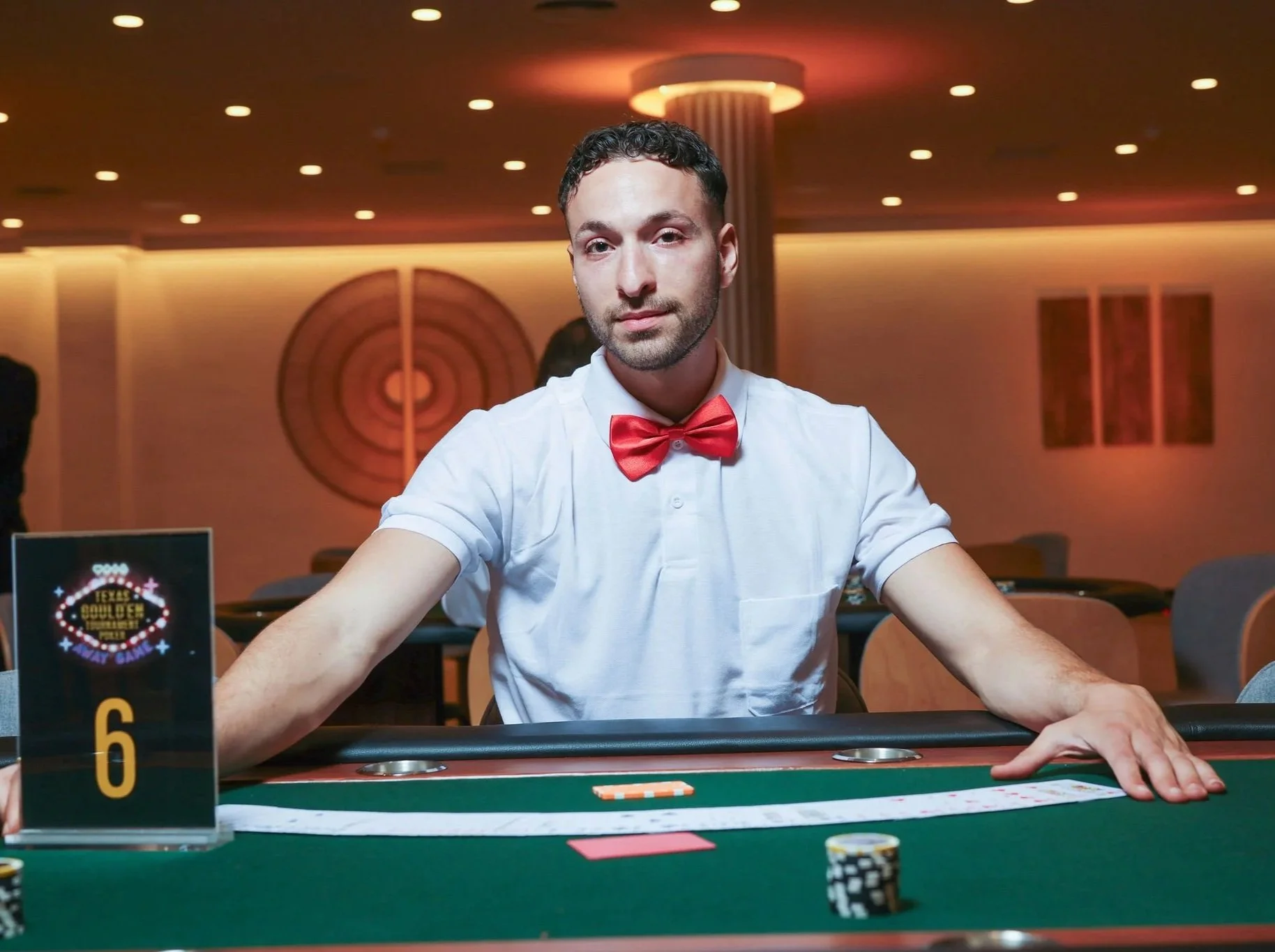 A man in a white shirt and red bow tie sitting at a poker table in a casino, with poker chips and cards in front of him.  The background features a warm, lit room with circular ceiling lights and decorative wall art at Anantara Villa Padierna Resort 
