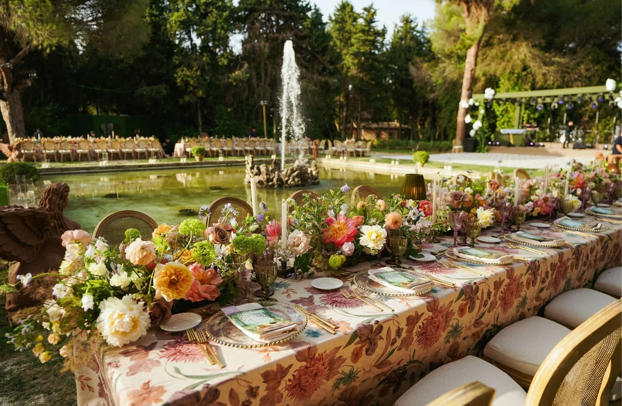 Decorated outdoor wedding reception with long floral table, chairs, and fountain in the background, surrounded by trees.