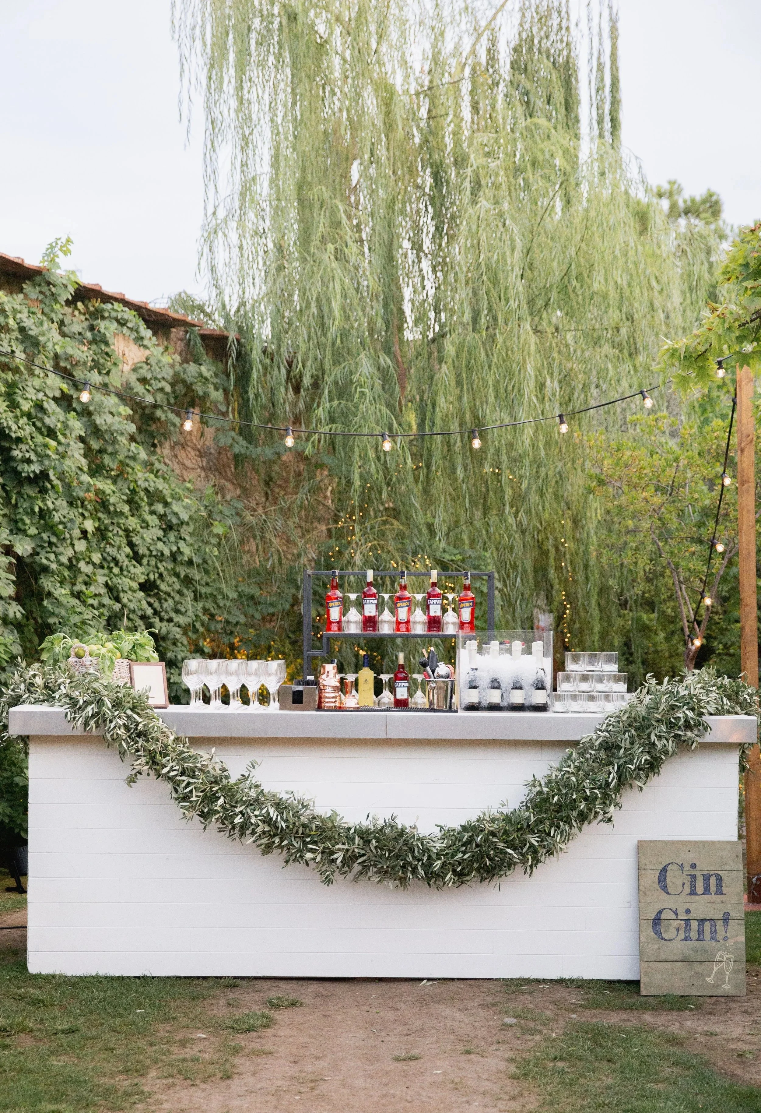 Outdoor bar setup with a white counter decorated with greenery, string lights overhead, various bottles of alcohol, glasses, and bar supplies, surrounded by trees and foliage, with a sign on the right that reads 'Gin Gin!'.