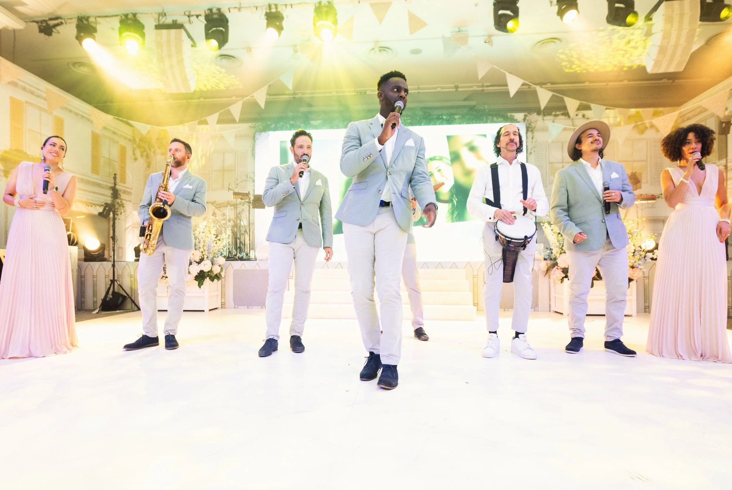Group of six performers on stage, four men and two women, dressed in light-colored formal attire, holding microphones and musical instruments, with colorful stage lighting and decorative banners overhead, performing in at Grosvenor House on Park Lane