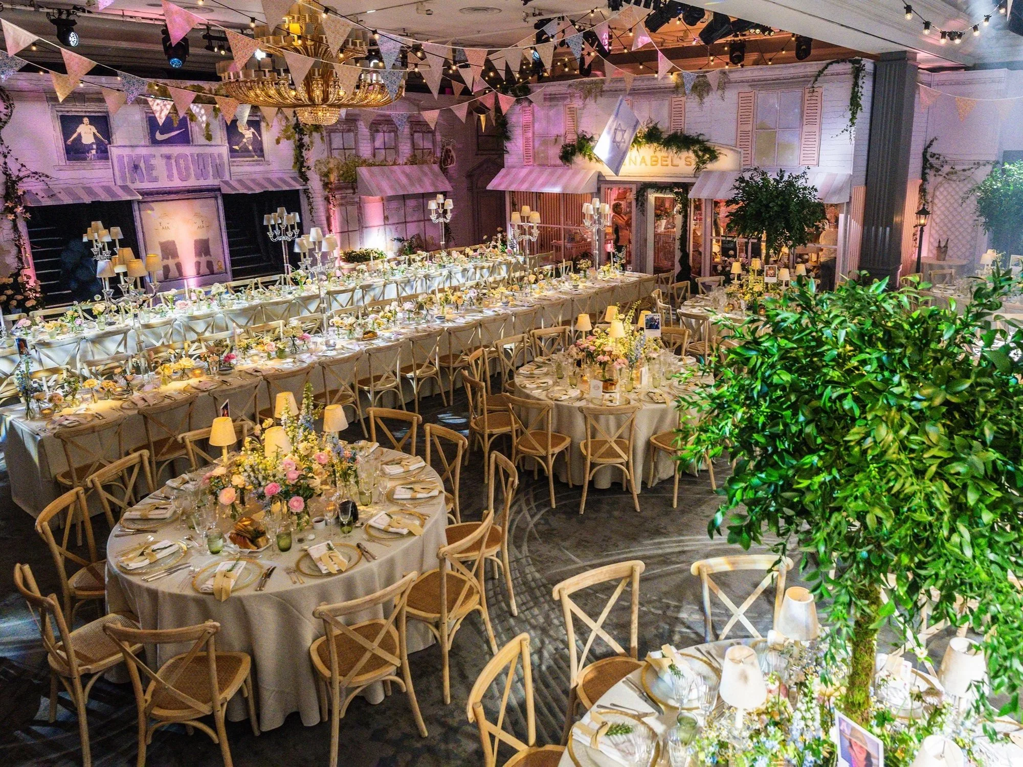 Decorated banquet hall with round tables, floral centerpieces, and elegant table settings. Wall and ceiling are adorned with bunting, string lights, and flags, including a visible Israeli flag. There are framed sports posters, a chandelier, and green