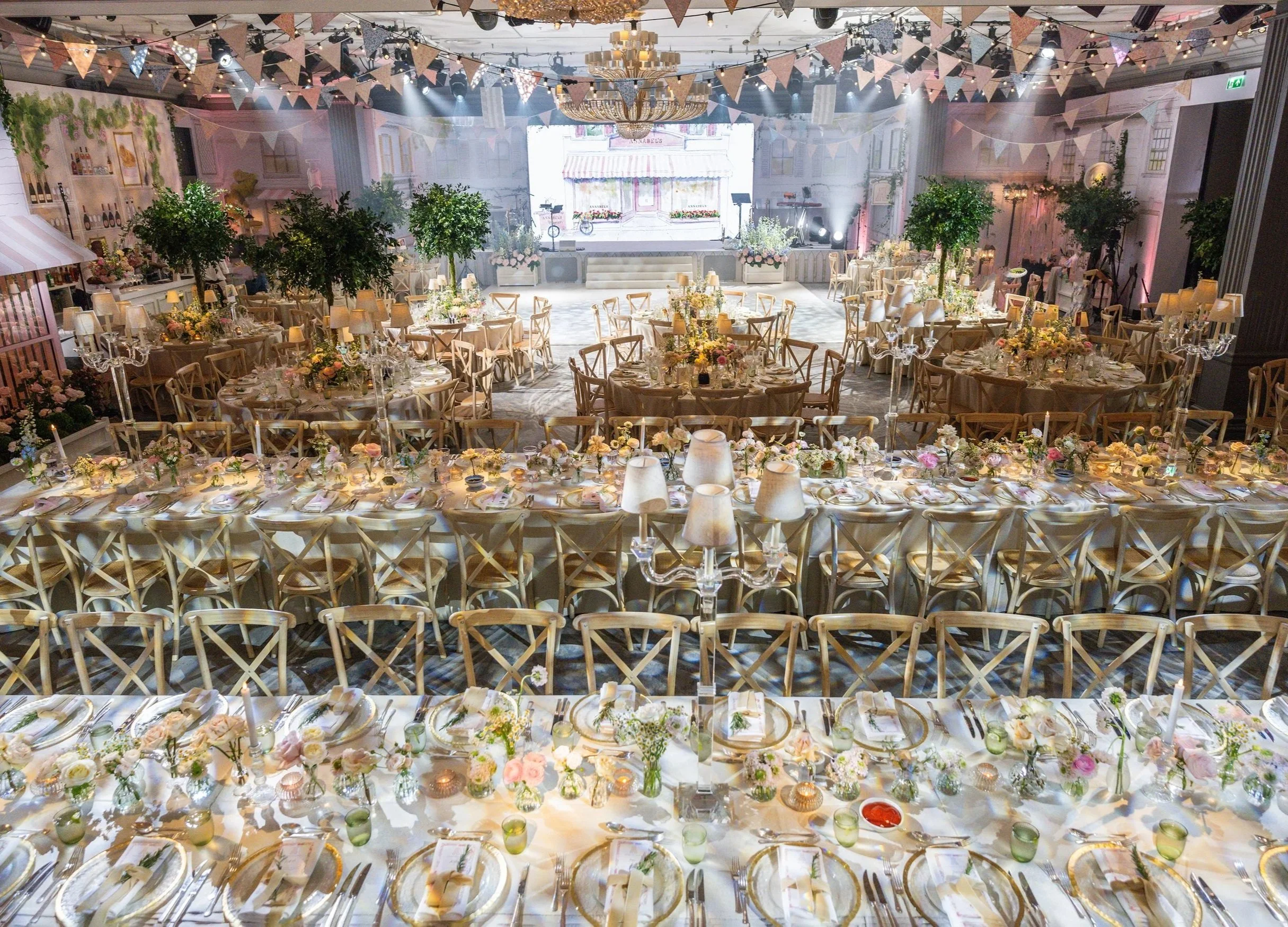 Decorated Grosvenor House on Park Lane with long banquet tables, floral centerpieces, candles, and set dining ware, with a stage at the back, hanging bunting, and indoor plants.