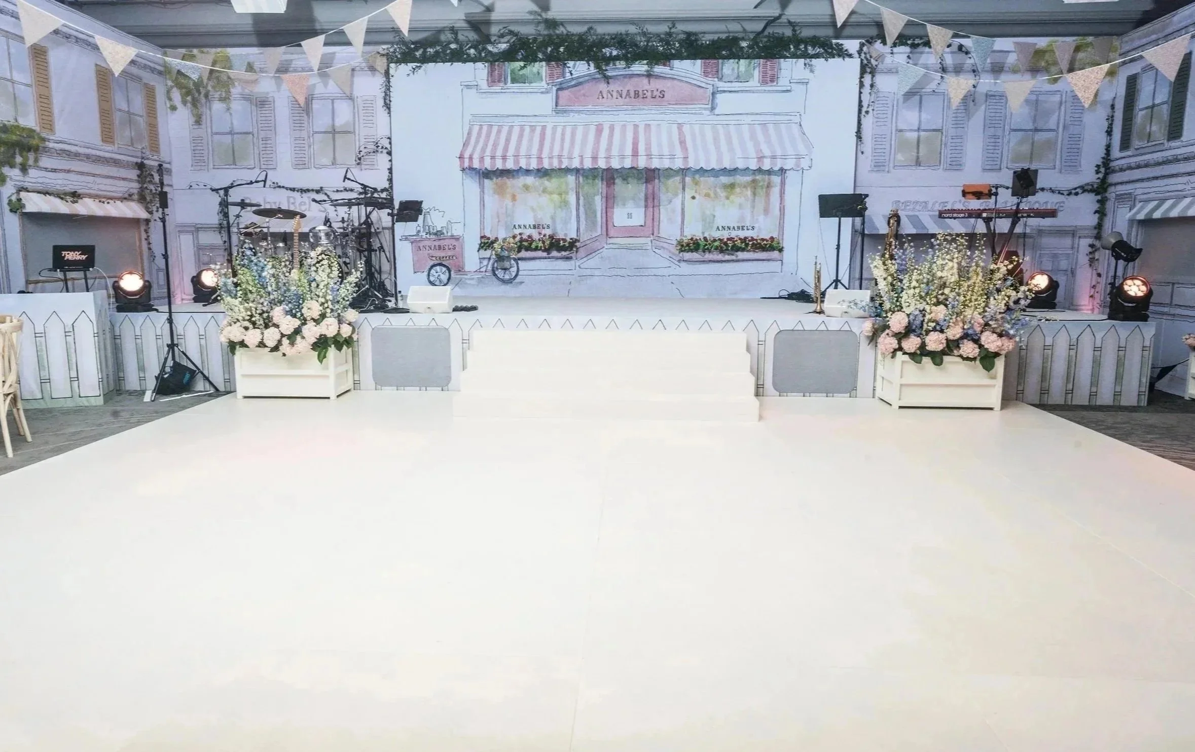 Event stage with a backdrop featuring a illustrated storefront, floral arrangements, stage lighting, musical instruments, and decorative bunting.