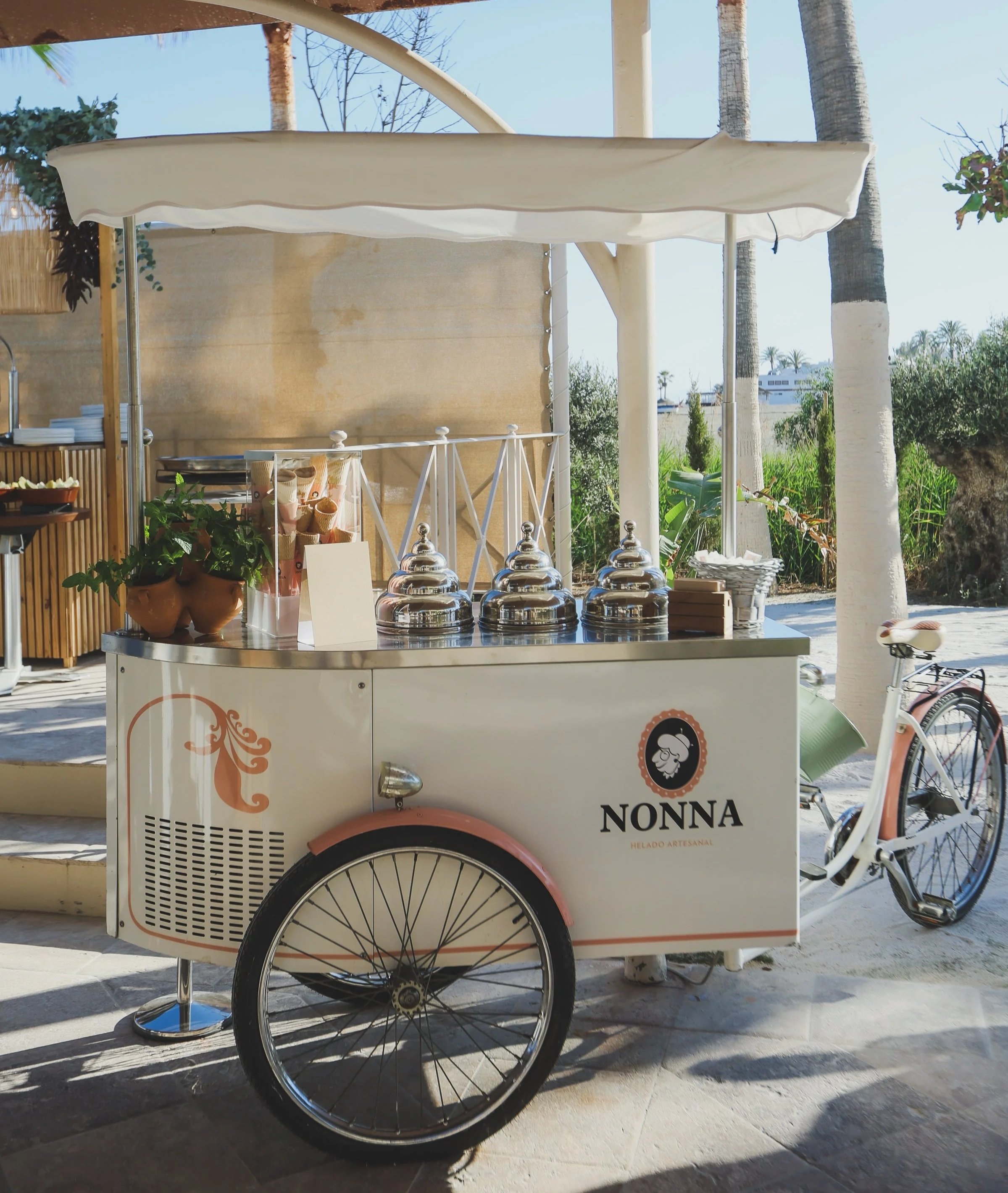 A white ice cream cart with black wheels and orange accents, branded with the name 'NONNA'. It is situated outdoors with trees and a fence in the background, and various ice cream containers are on the cart.