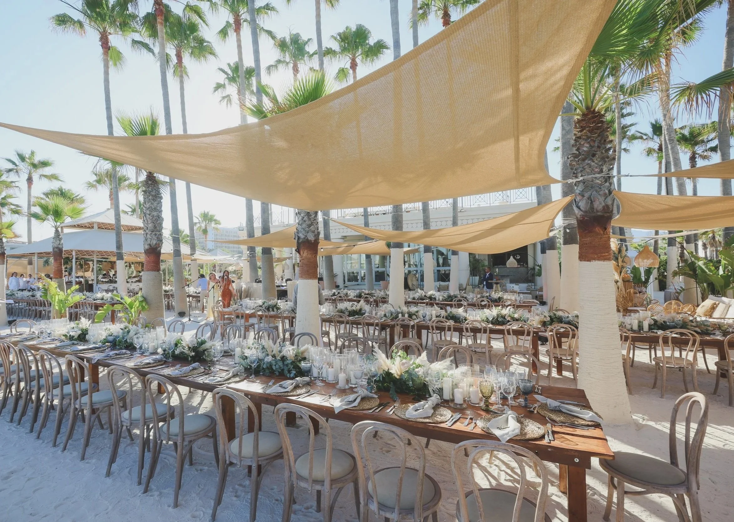 A beachside outdoor event setup with long tables decorated with white and blush floral arrangements, place settings, and glassware. Beige sail shades are stretched overhead for shade, and tall palm trees surround the area, creating a tropical atmosphere.