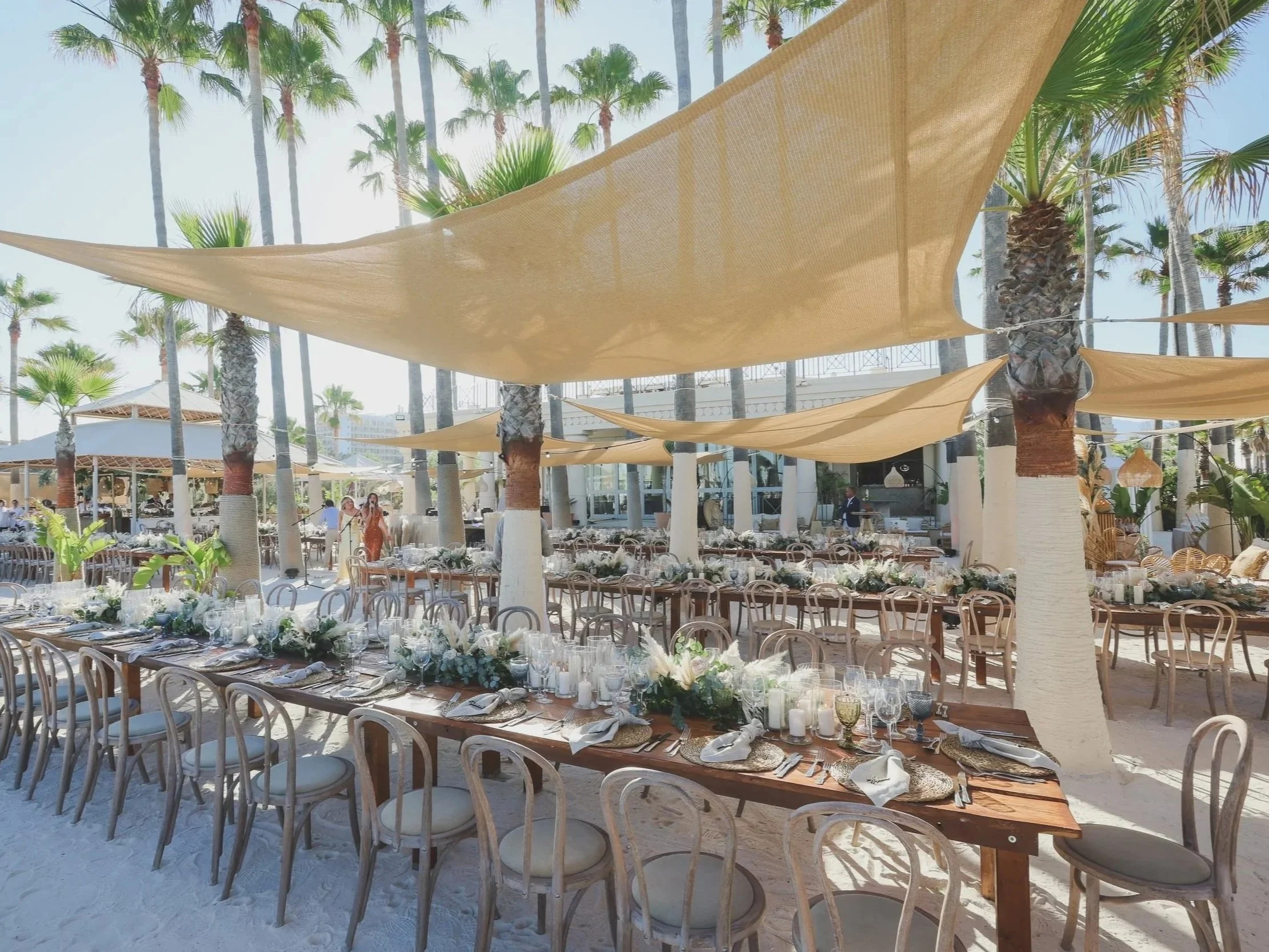 Wedding reception setup outdoors on a beach with long tables decorated with white flowers, candles, and glassware, surrounded by palm trees and shade sails.