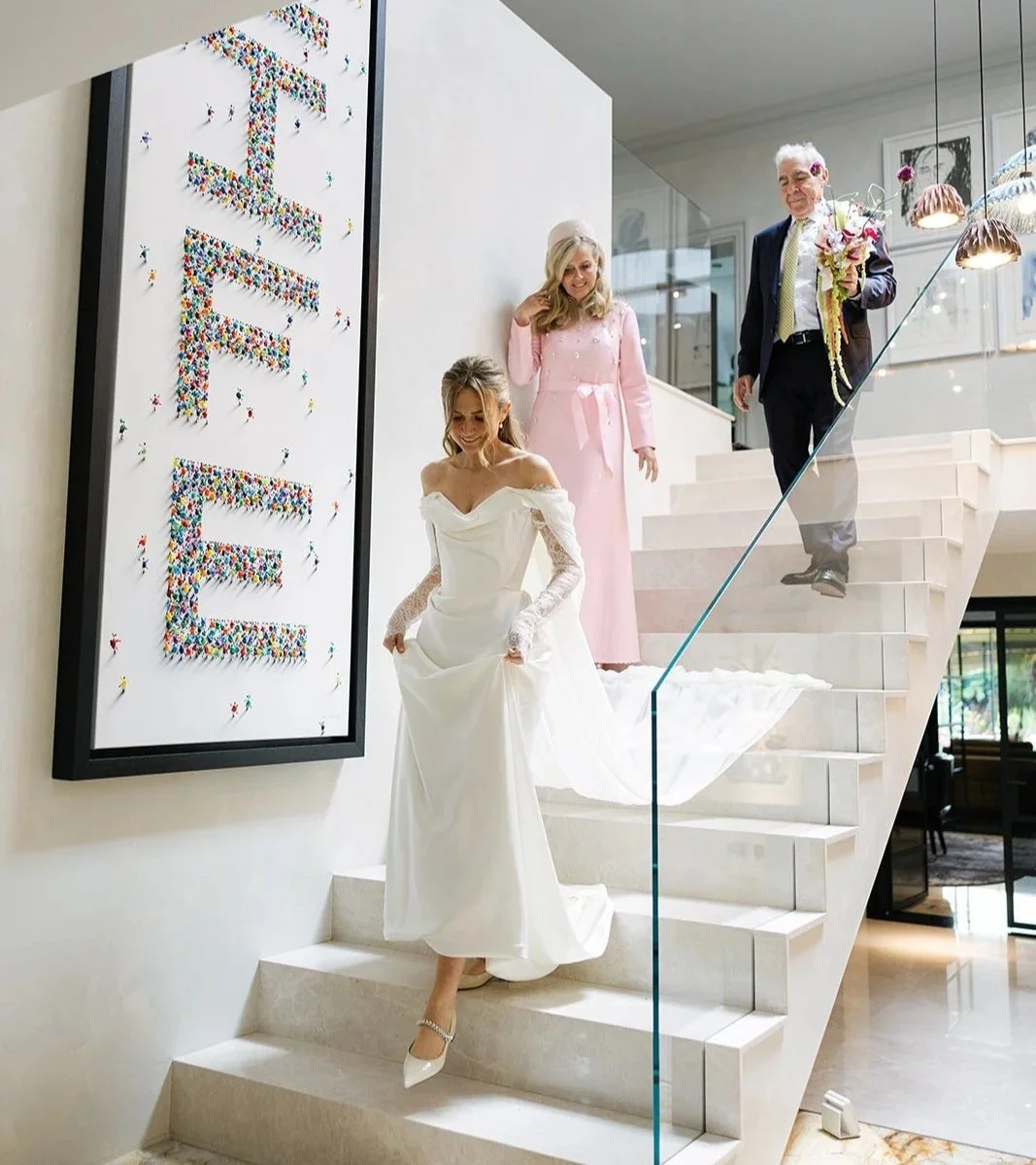 A bride in a white wedding dress with lace sleeves descending a staircase, accompanied by a woman in a pink dress and an older man in a suit holding a bouquet, inside a modern building with a large colorful artwork on the wall.