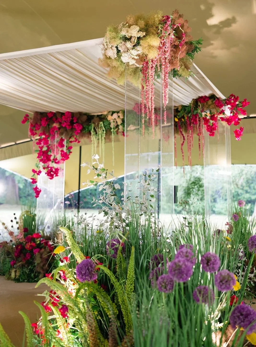 Decorative chuupah floral arrangement with pink, purple, and green flowers hanging from a ceiling with beige drapery, set against a backdrop of greenery.
