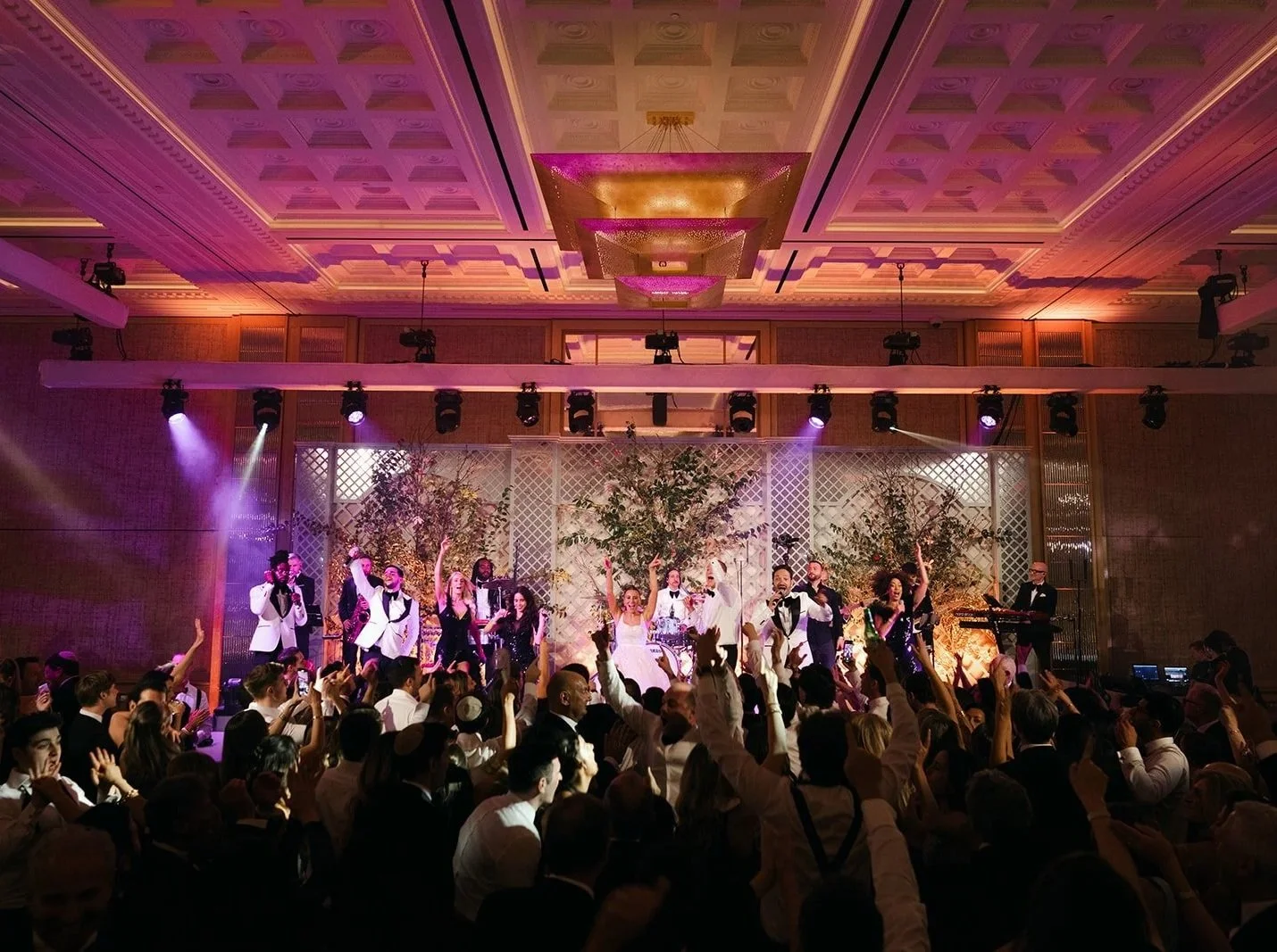 Group of performers on stage singing and dancing with audience raising hands in celebration, in a ballroom with decorative trees and lighting at The Peninsula London