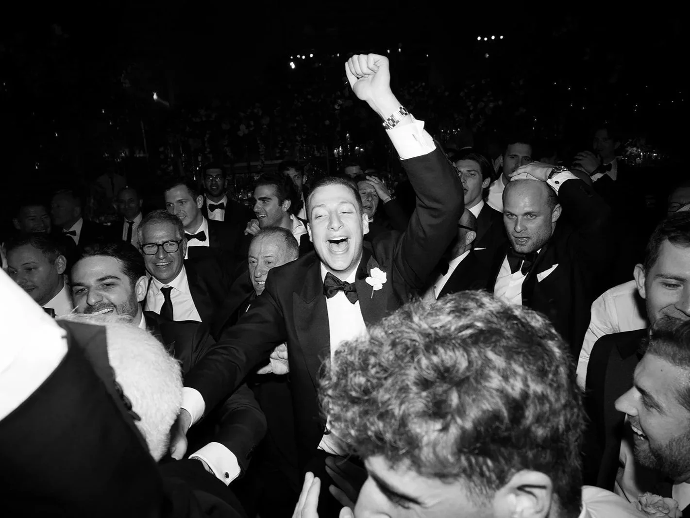 A group of men and groom in tuxedos at a celebration, with one man raising his hand and smiling excitedly, in a black and white photograph at The Peninsula London