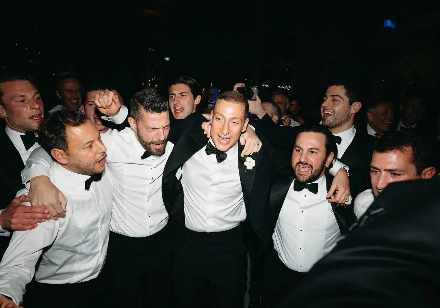 A group of men in tuxedos and bow ties celebrating together at wedding party, the groom in the center being embraced by friends, at The Peninsula London Hotel