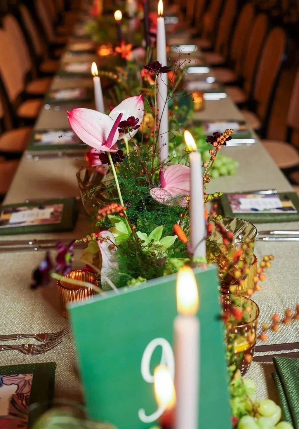 Close-up view of a creative wedding dinner table centerpiece with candles, pink anthuriums, and various greenery and small decorative flowers, set on a beige tablecloth. Background shows chairs and table settings.