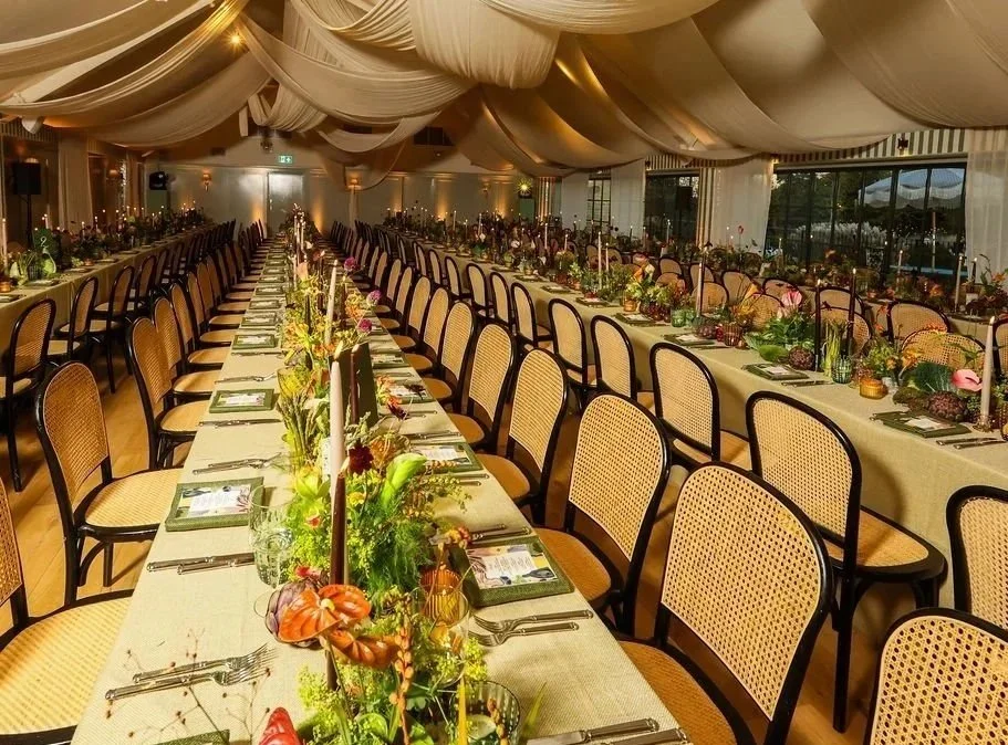 Long banquet tables set with floral centerpieces, candles, and place settings inside a decorated tent for a formal wedding reception.