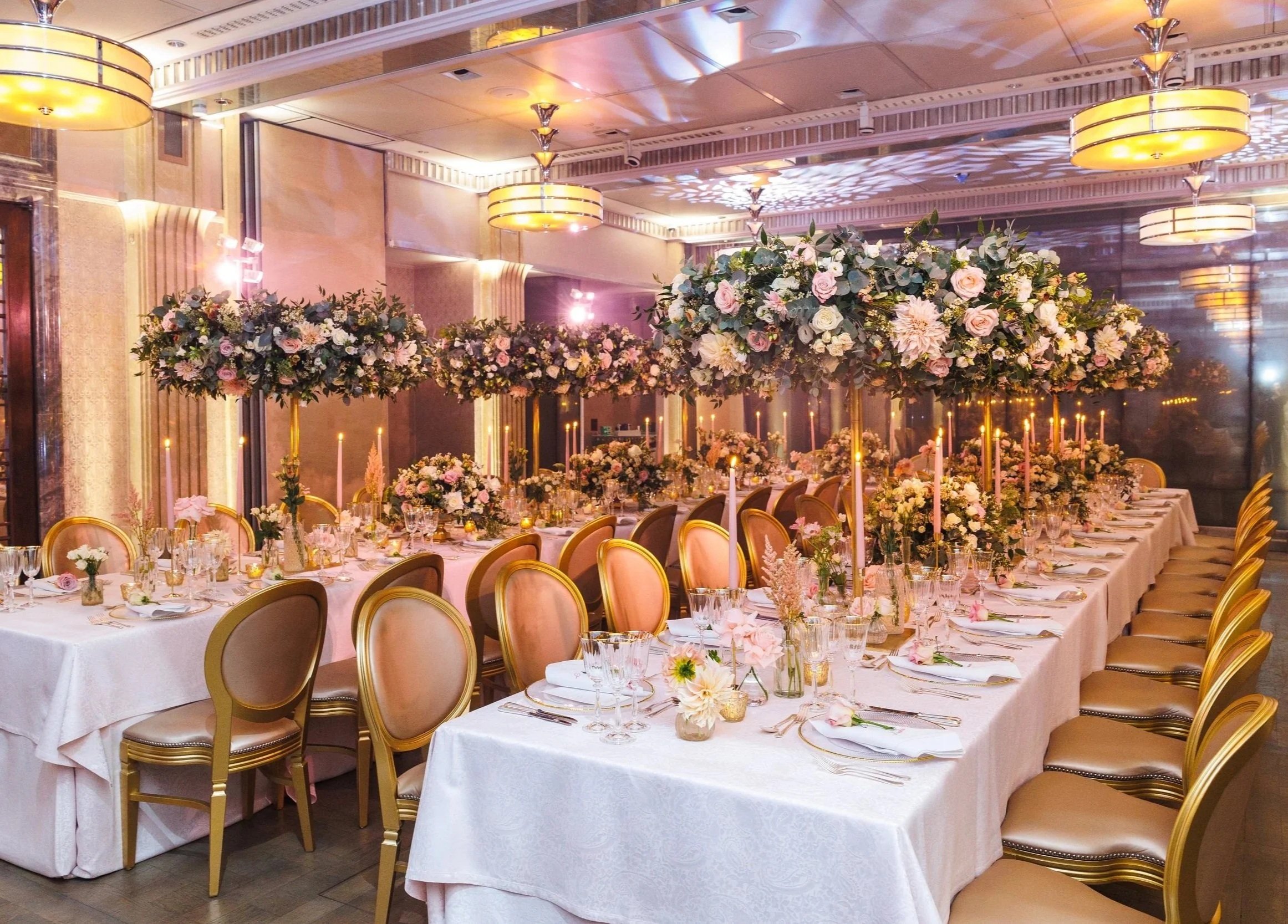 Elegant banquet hall decorated with pastel-colored flowers and candles on round and rectangular tables, with chairs arranged for a formal event.