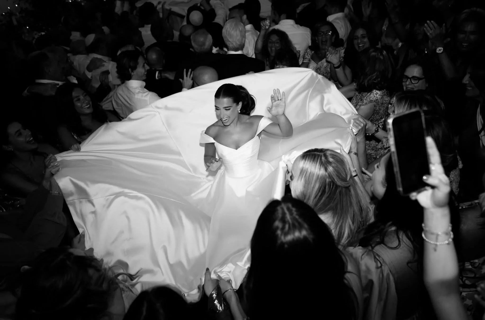 A bride in a white wedding dress with an off-the-shoulder neckline smiling and waving among a crowd of people at a wedding reception.