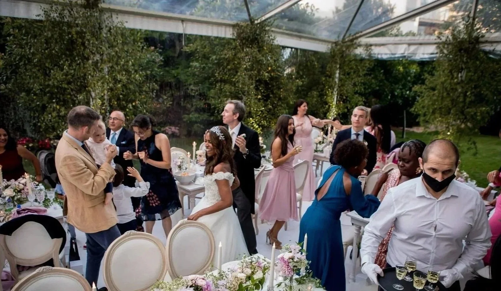 People celebrating at a wedding reception in a greenhouse with floral decorations and tables set for a meal.