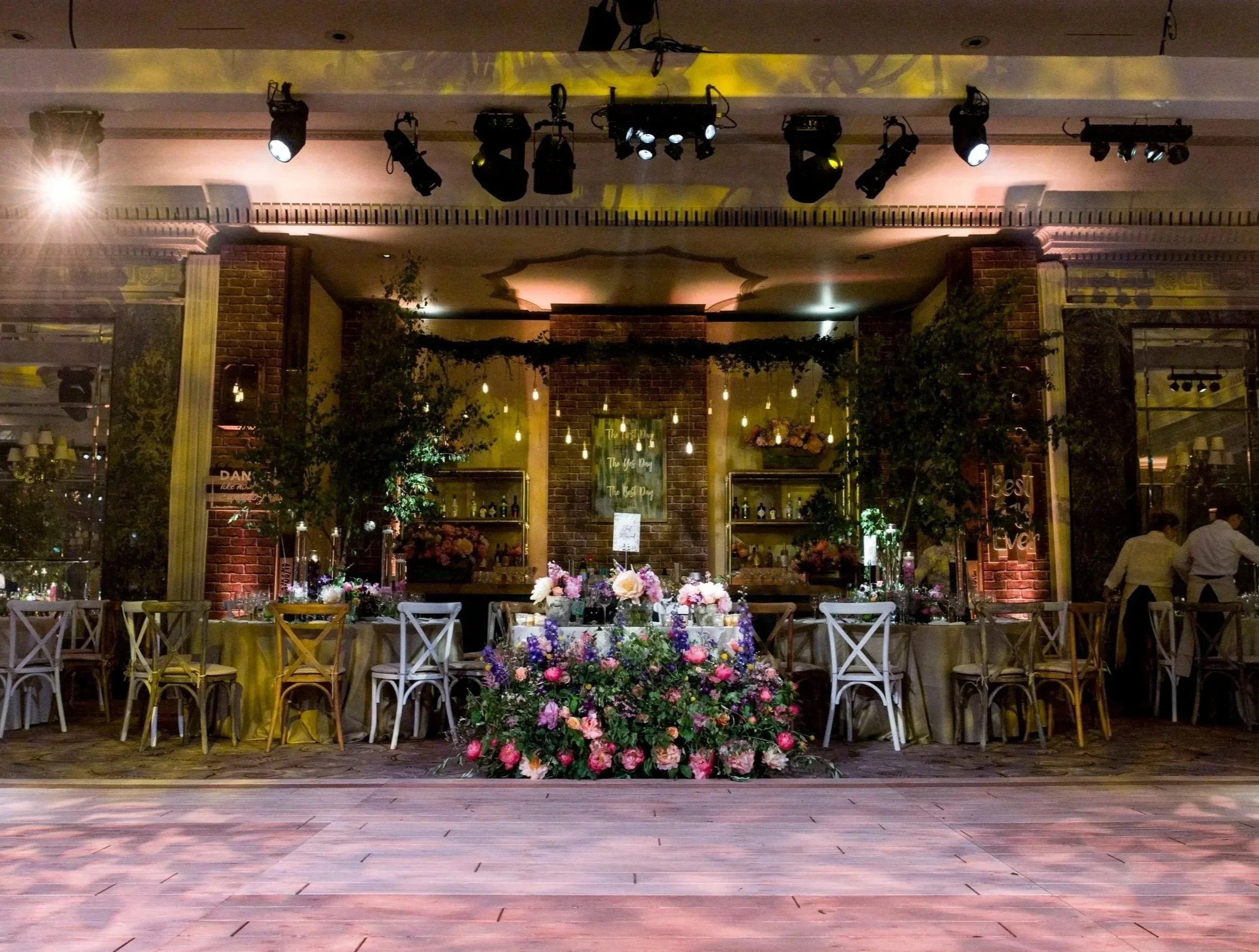 The image shows an indoor event space decorated for a celebration, with a large floral arrangement on the floor in the foreground, a row of chairs along the front, and a backdrop featuring brick columns and greenery. There are tables with flowers and