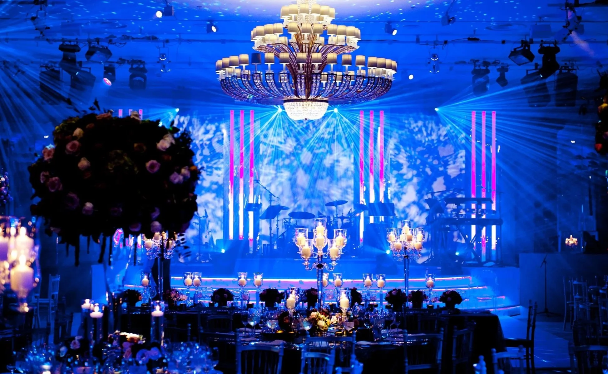Elegant banquet hall with a large chandelier, colorful lighting effects, flower arrangements, and tables set for a wedding at Grosvenor House