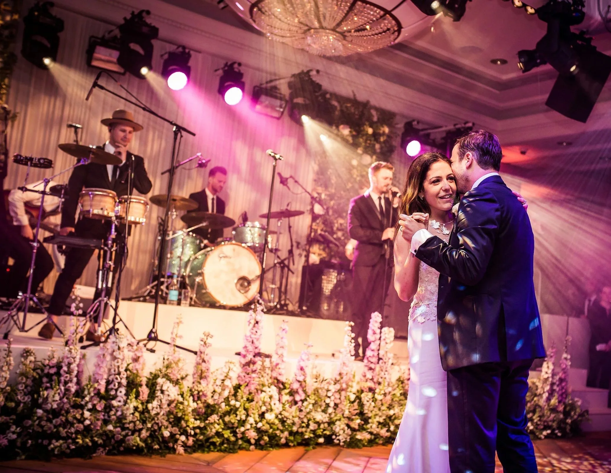 A bride and groom dance closely at their wedding reception, with a live band performing on stage behind them. The stage is decorated with pink flowers and draped with light-colored fabric at Rosewood London