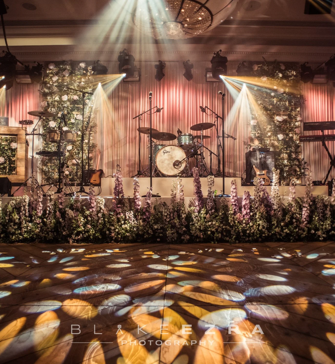Stage decorated with pastel pink curtains, surrounded by floral arrangements, with musical instruments including drums, guitars, and microphones, lit by stage lights and spotlight reflections on the wooden floor. Rosewood London