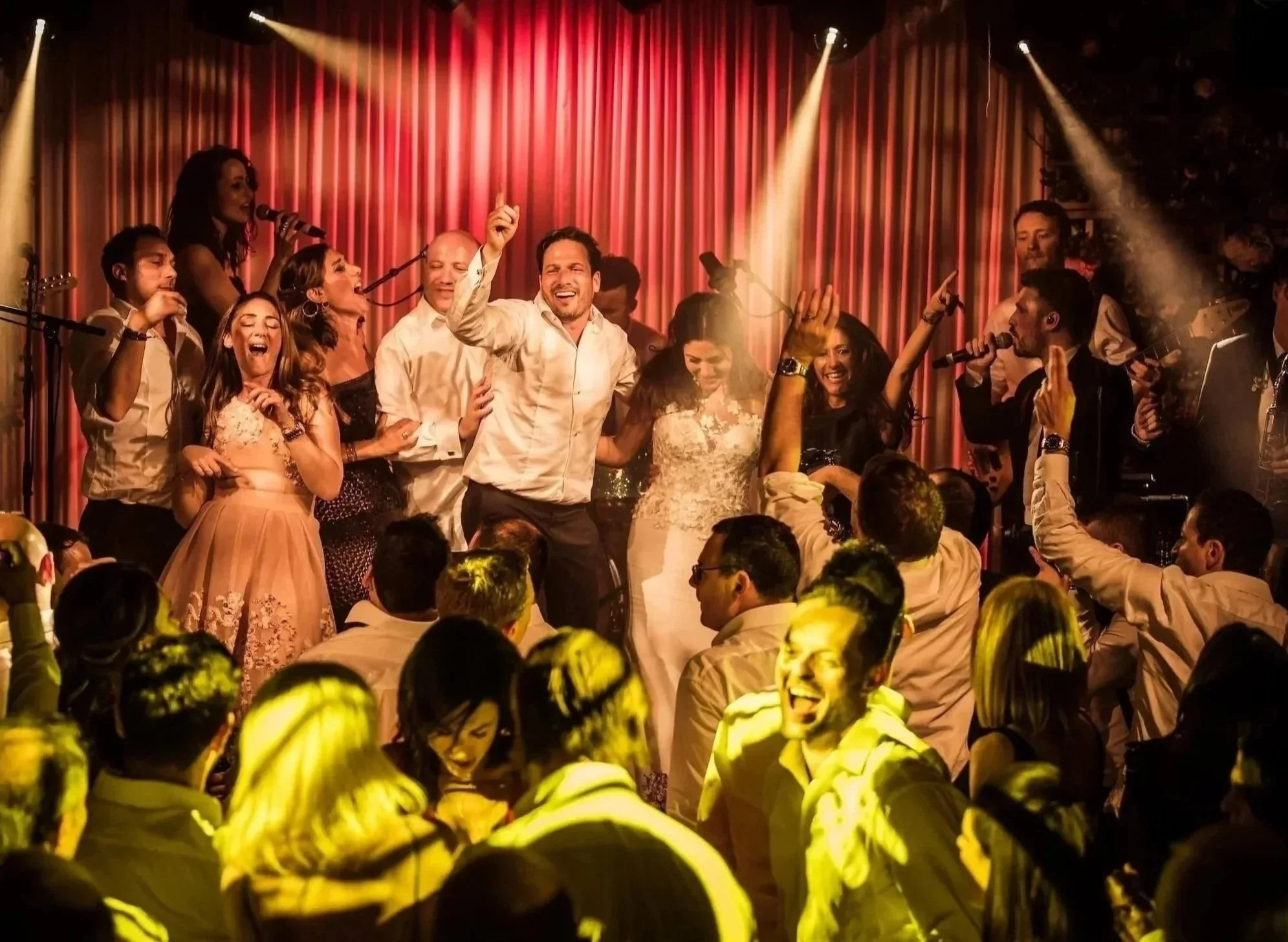 People dancing and singing on stage with a red curtain background, illuminated by stage lights, at a wedding at Rosewood London