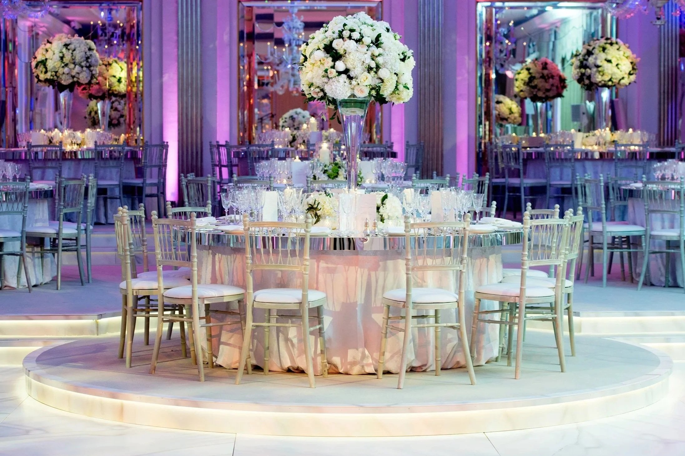 Elegant event venue decorated with large floral centerpieces featuring white roses and hydrangeas on tall glass vases, surrounded by tables set with glassware, candles, and white chairs, illuminated with colorful purple, pink, and blue lighting.