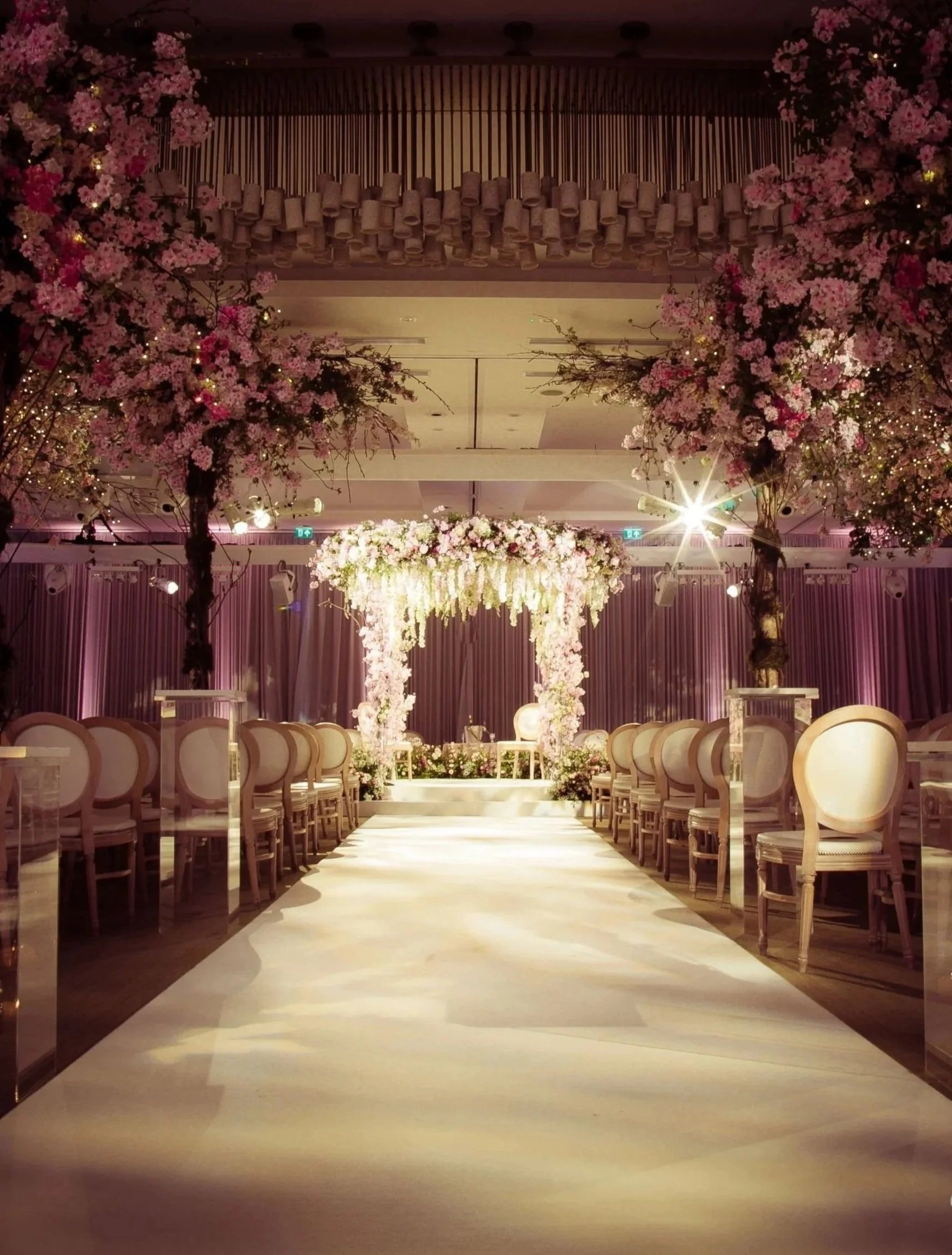 Elegant wedding ceremony setup with floral arch, pink and white cherry blossom trees, and rows of chairs on either side, in a decorated ballroom at Rosewood London