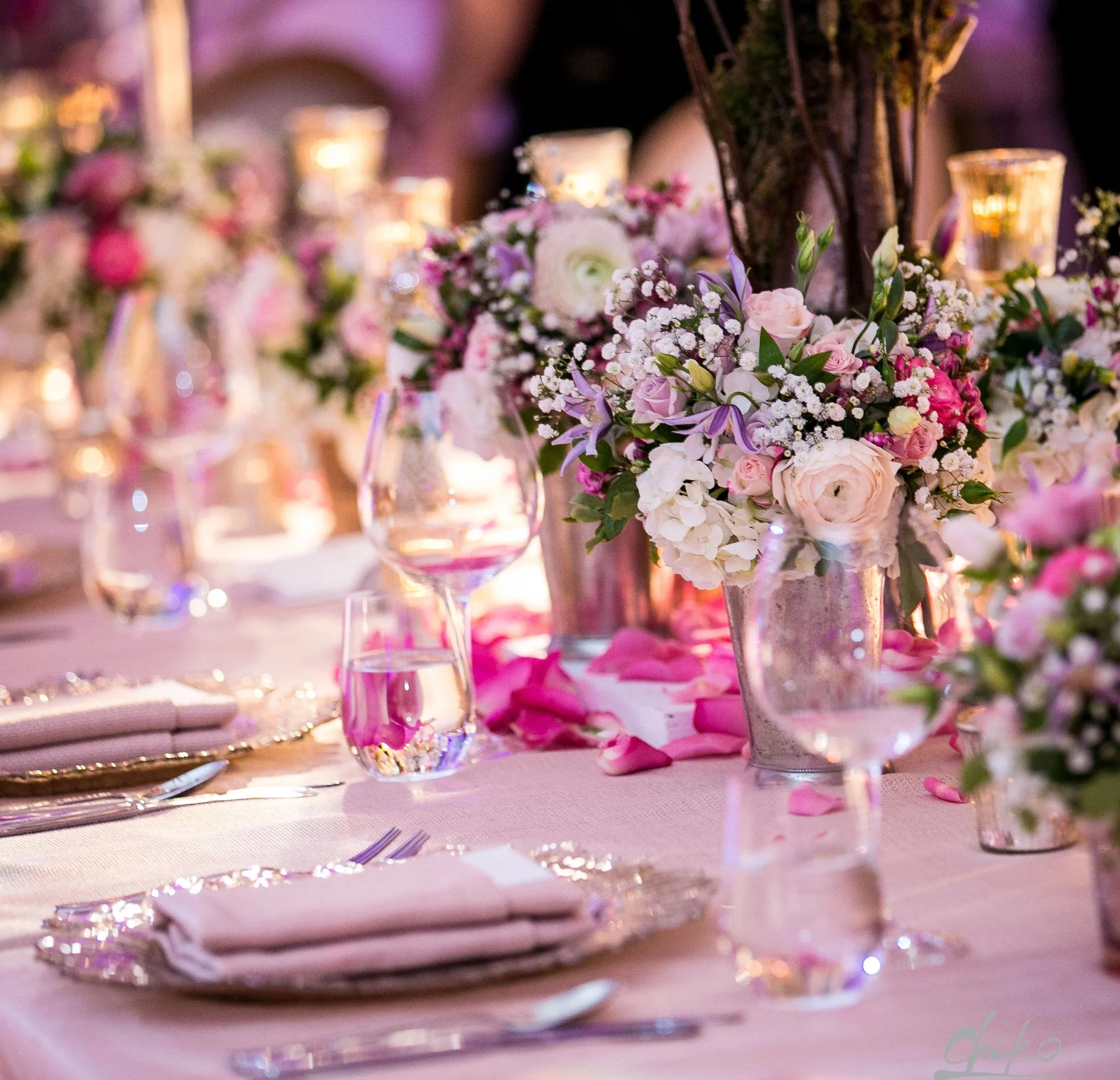 A decorated table with pink and white floral centerpieces, candles, glassware, and pink rose petals, suggesting a formal event or celebration.