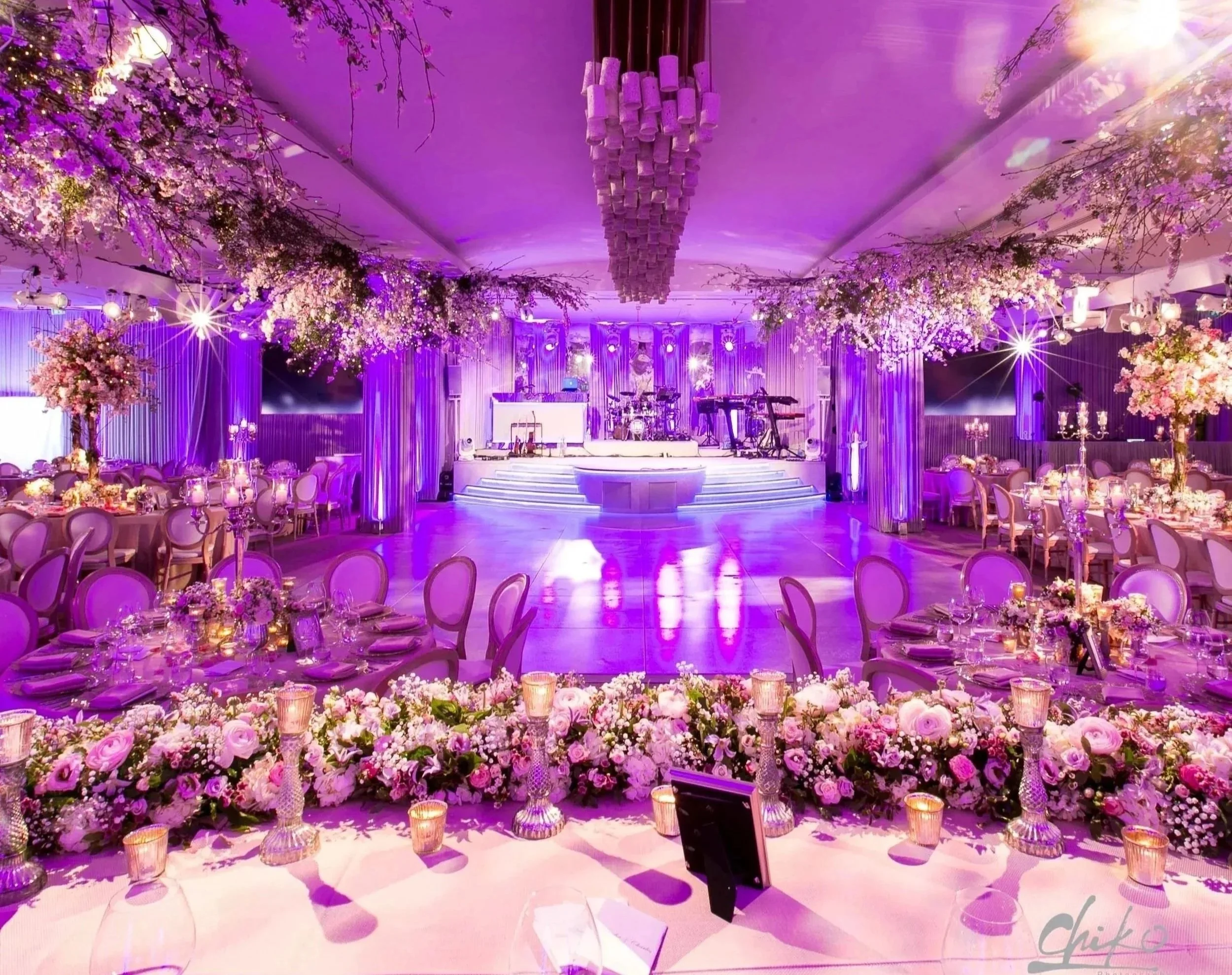 Elegant event hall decorated with purple and pink flowers, a staircase in front of a stage with musical instruments, floral arrangements, candles, and purple lighting.