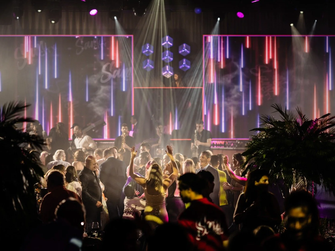 Crowd dancing and mingling at a lively indoor concert or nightclub with a large LED display screen and several performers playing music on stage, illuminated by colorful lights Bar Mitzvah