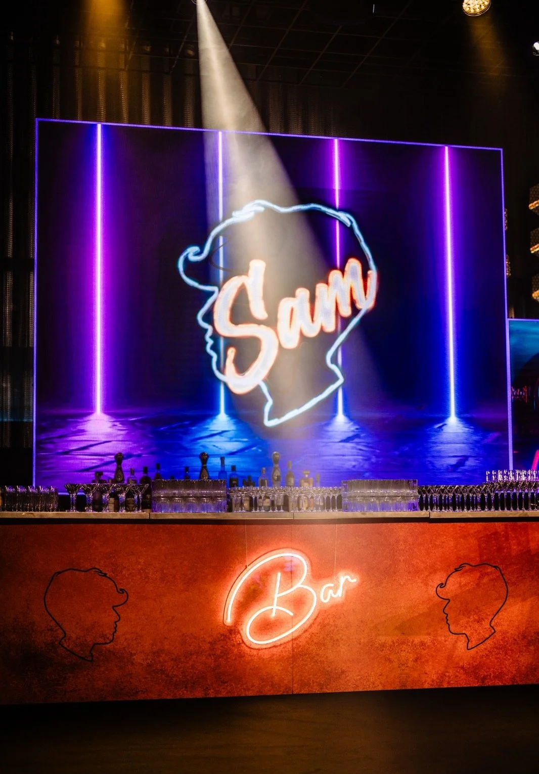 A bar with neon signs, including a large neon sign of a woman's profile with the name 'Sam' inside it, and the word 'Bar' below. The bar has rows of glassware and bottles, with purple and blue lighting Bar Mitzvah