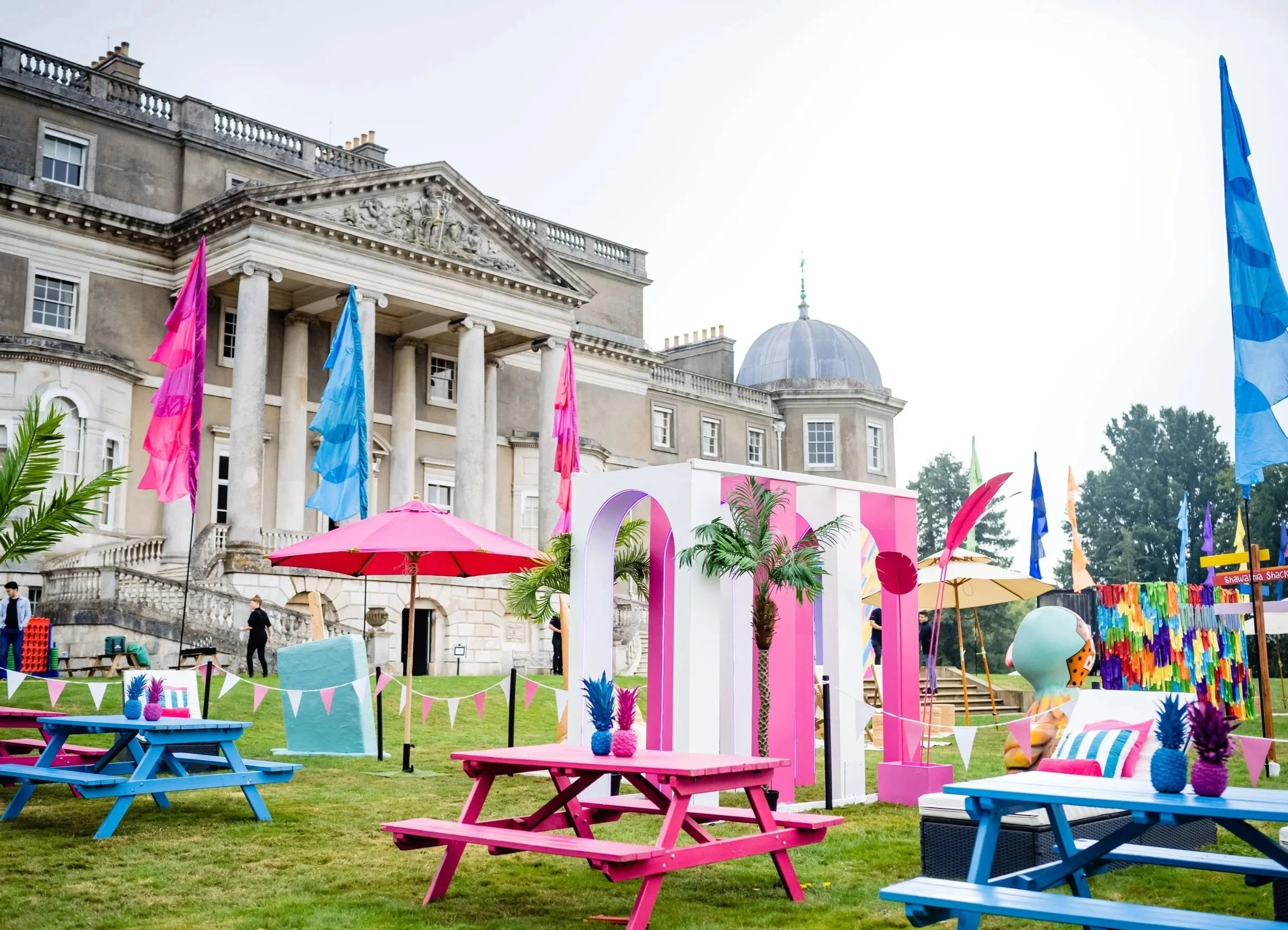 Colorful outdoor event setup featuring pink and blue picnic tables, umbrellas, tropical decorations, and a historic building in the background ata  Bar Mitzvah at  Wrotham Park