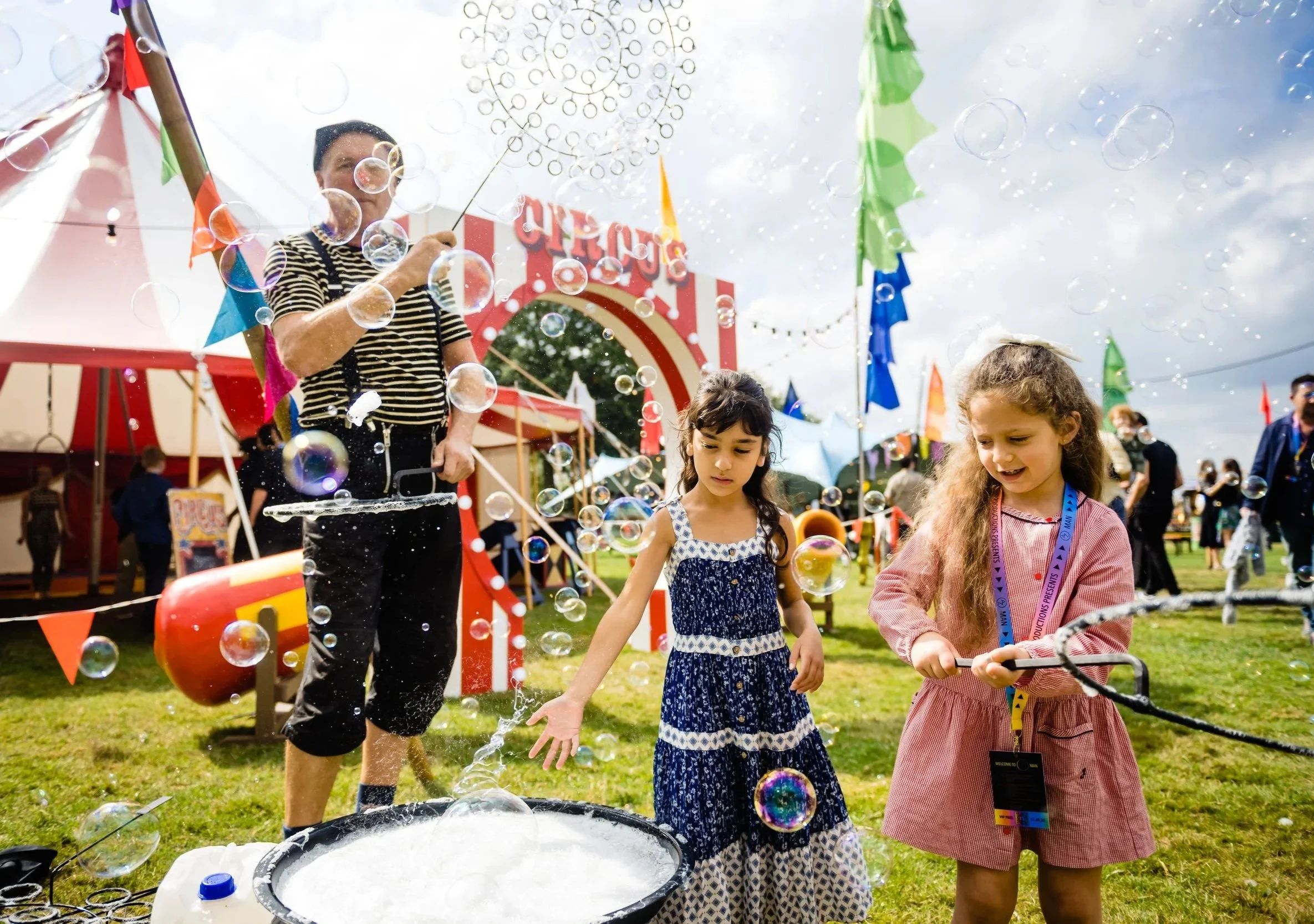 Three young girls and a woman are at an outdoor festival, playing with bubbles. The woman, dressed casually and wearing a striped shirt, is using a wand to create bubbles. The girls are observing and engaging with the bubbles, appearing happy and cur