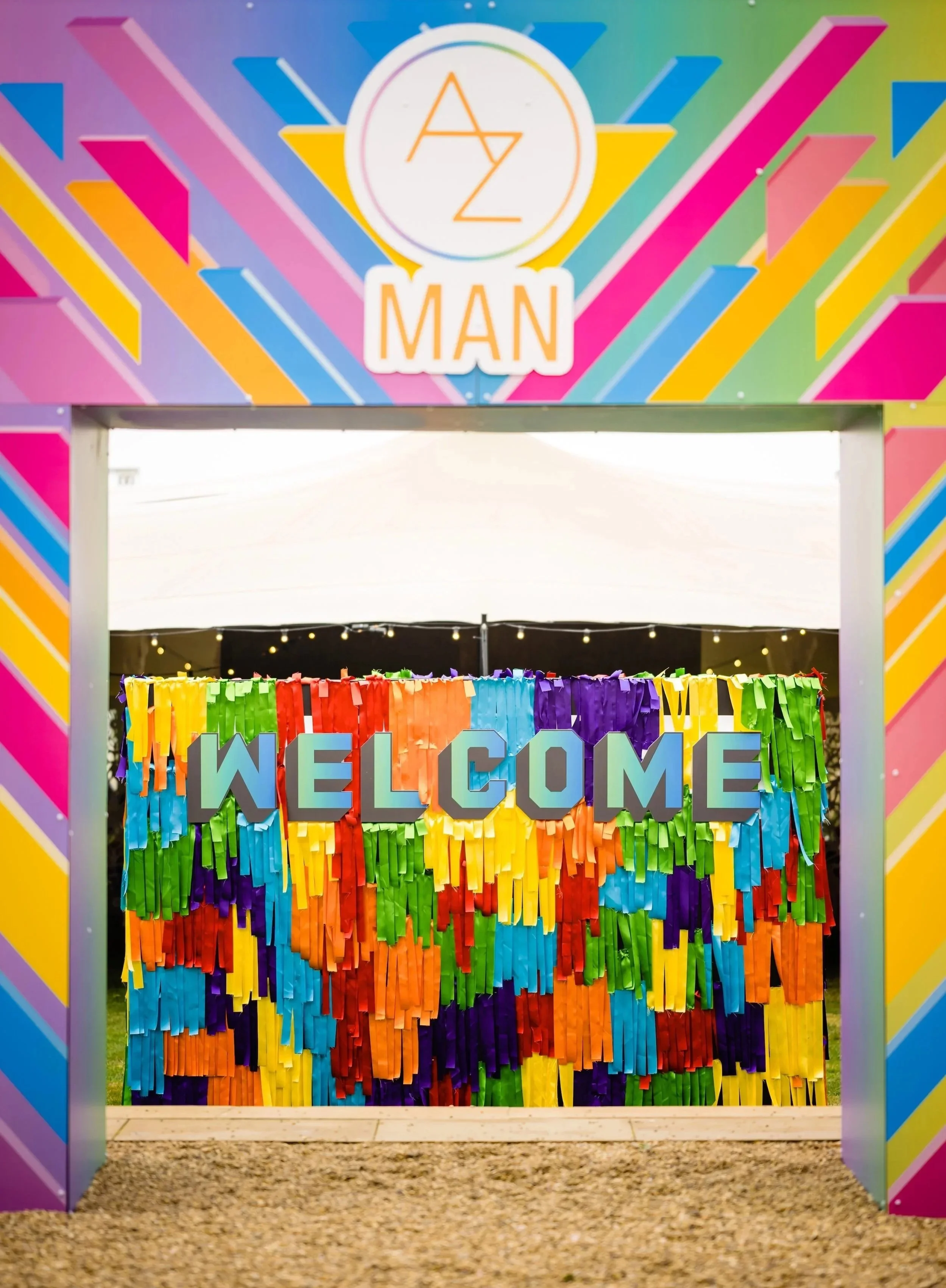 An entrance decorated with a colorful rainbow-themed archway, a 'WELCOME' sign made of rainbow-colored paper strips, and a sign that reads 'AZ MAN' featuring a rainbow-colored circle and various geometric patterns for a Bar Mitzvah at  Wrotham Park