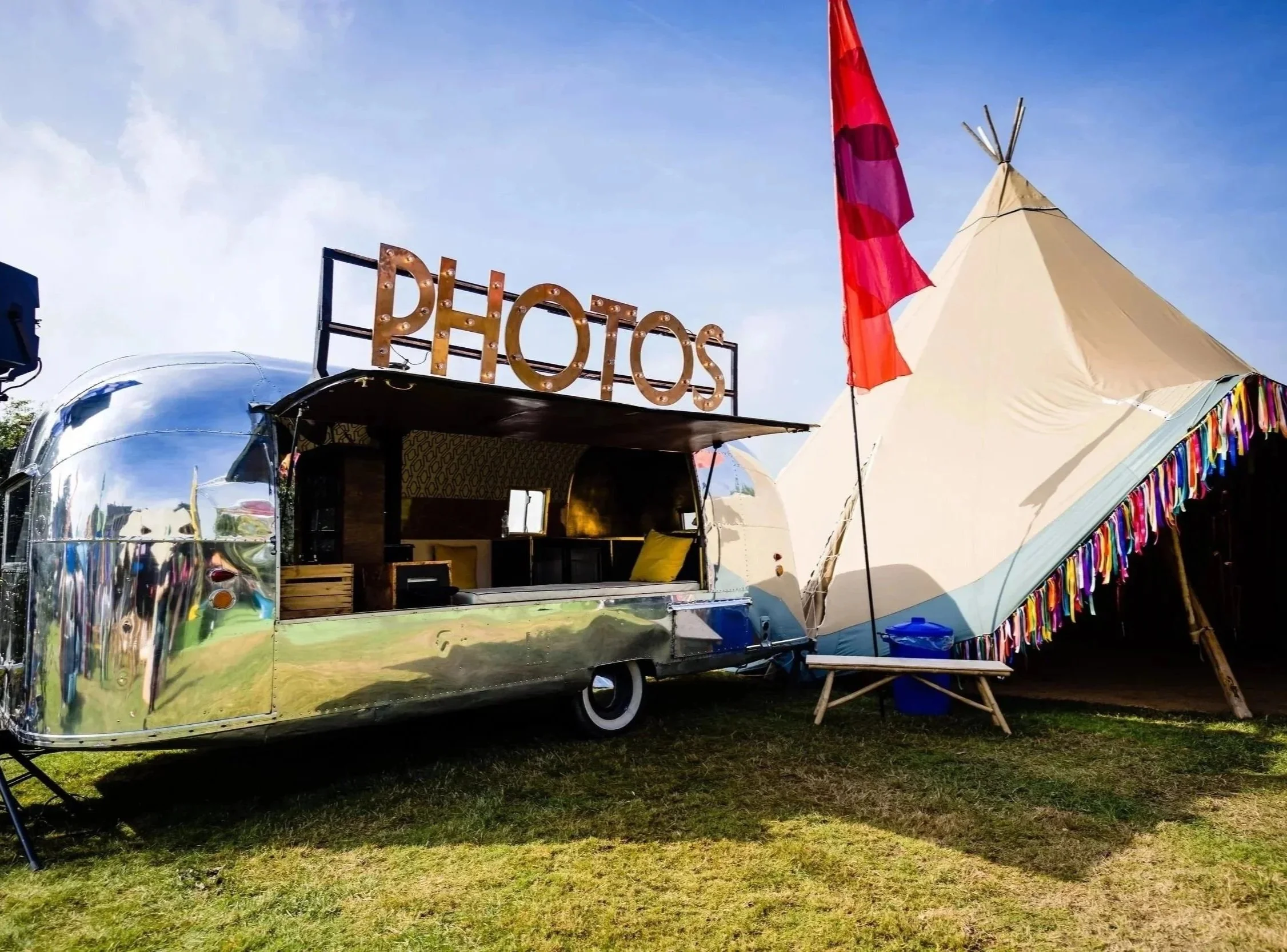 A shiny silver vintage trailer and a tent with colorful fringe. The trailer has a sign with glowing letters that says "PHOTOS."  at a Bar Mitzvah at  Wrotham Park