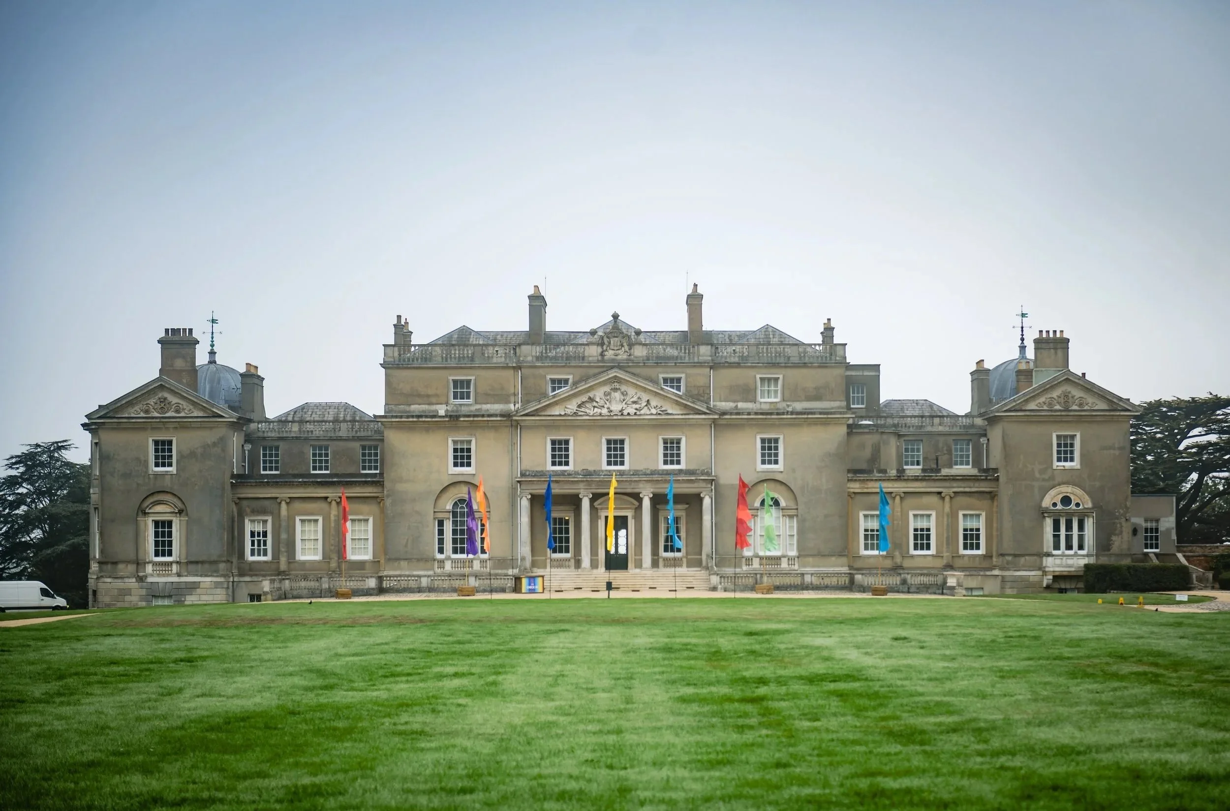A Bar Mitzvah at  Wrotham Parkwith a symmetrical facade, multiple chimneys, and decorative stone crestings on the roof. In front of the mansion, there is a well-maintained lawn with colorful flags hanging from the building's front. 