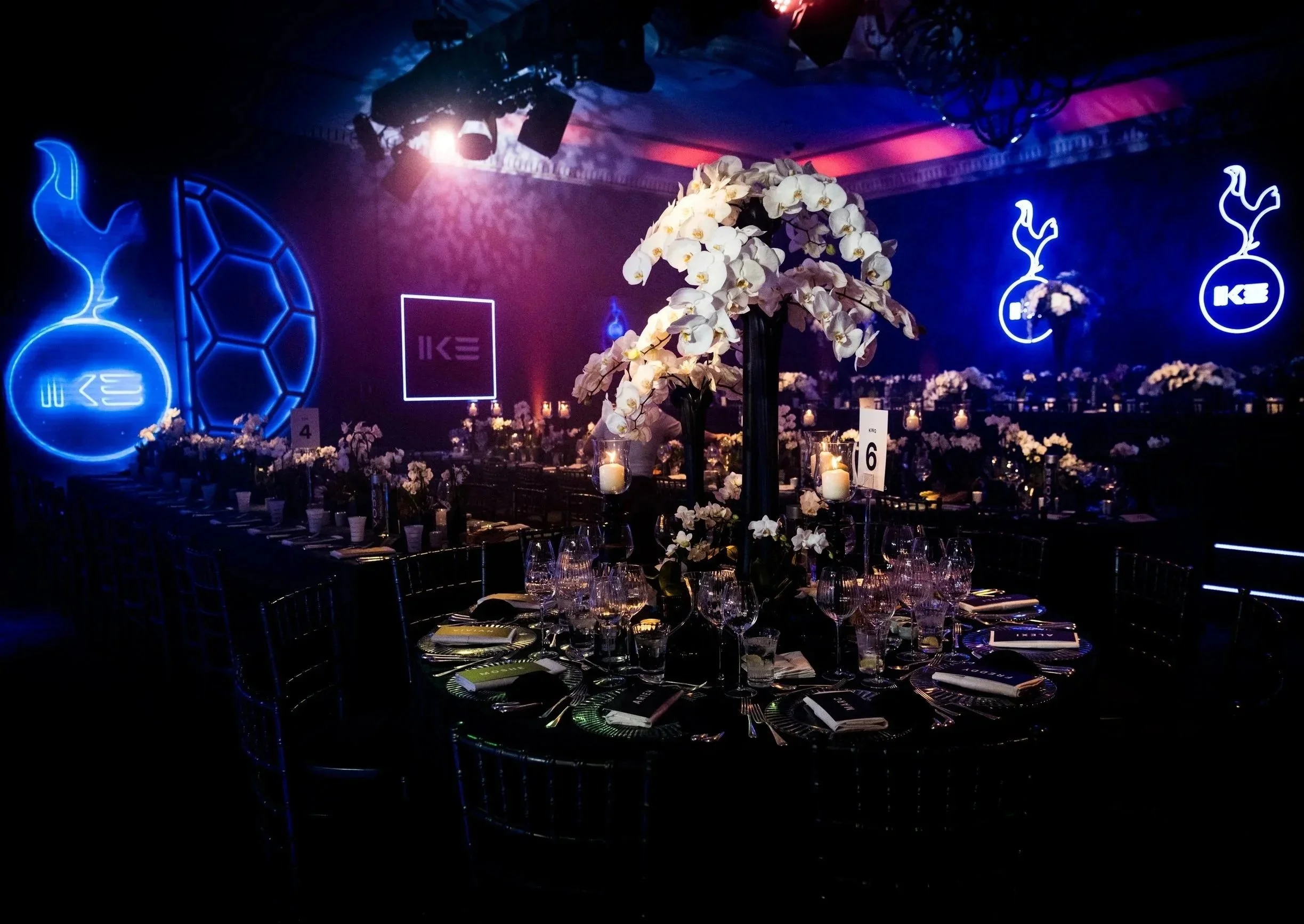 A banquet table decorated with white flowers in the center and multiple candles, set in an event space with dark walls illuminated by neon blue and purple lights for a Football themed luxury Bar Mitzvah