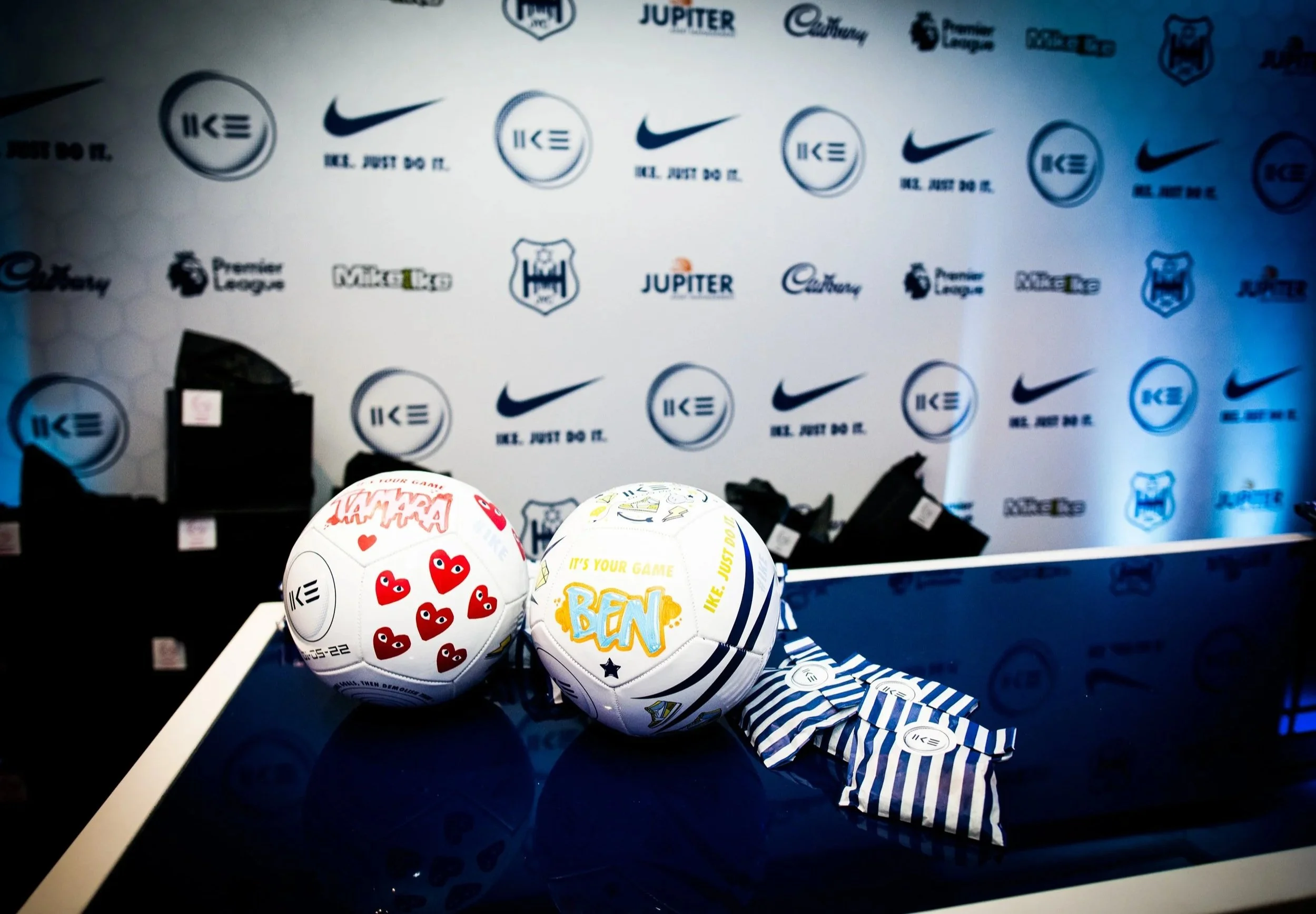 Several decorated soccer balls on a table with a backdrop featuring various logos and slogans like Nike, JUPITER, and 'WE. JUST DO IT' for a Football themed luxury Bar Mitzvah