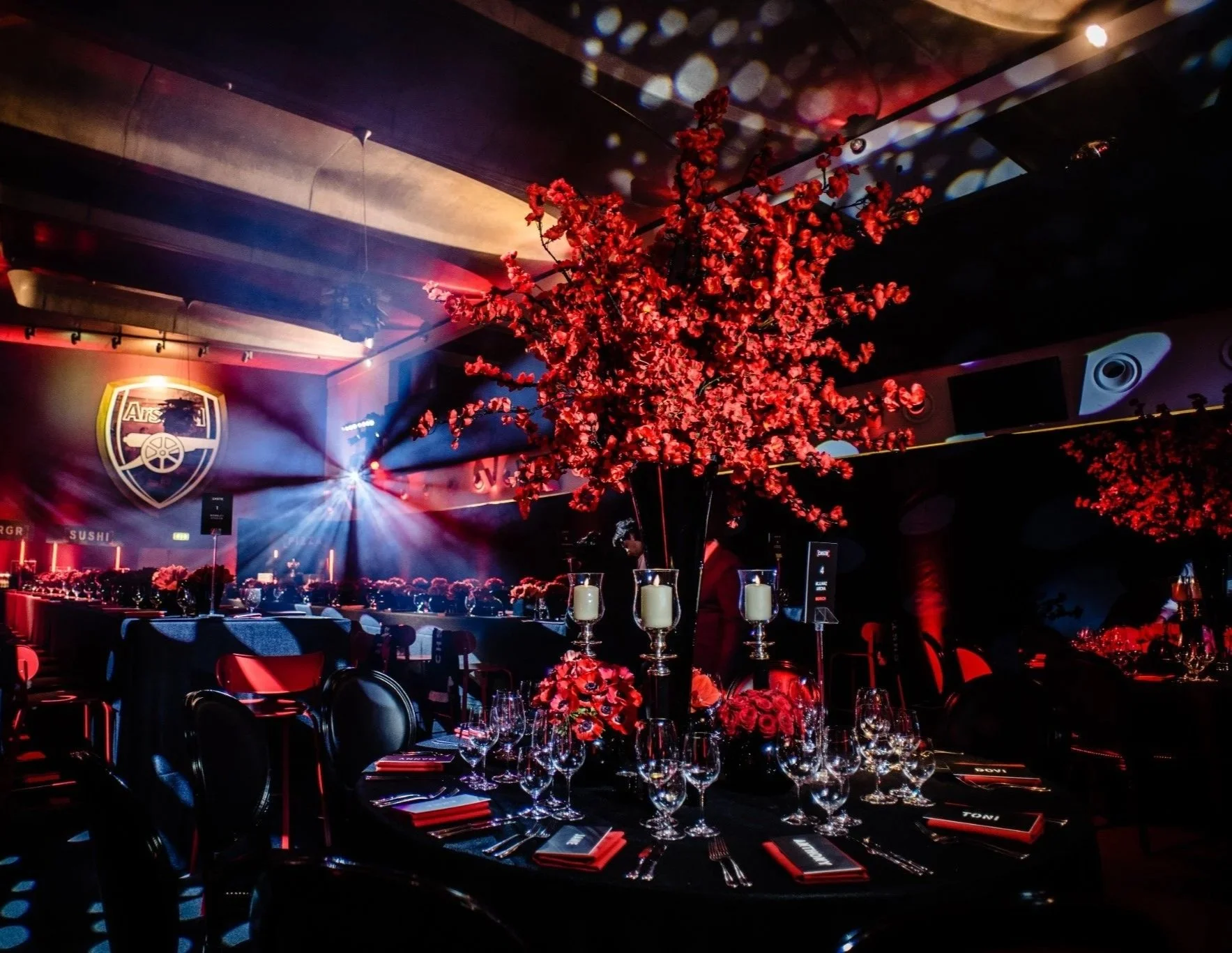 Indoor event space with a large centerpiece of red flowers on a black table, surrounded by wine glasses, plates, and candles, with a stage lit by colorful lights and an Arsenal logo on the wall.