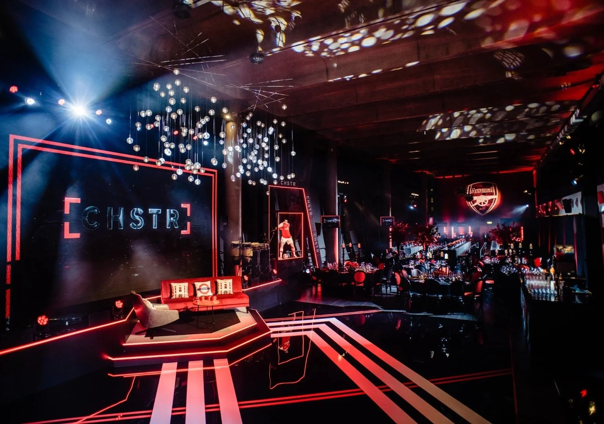 A large, dimly lit event space decorated with red and black accents and illuminated by colorful lighting. The space features a stage on the left with a seating area in front, a large screen displaying a logo with a cannon and 