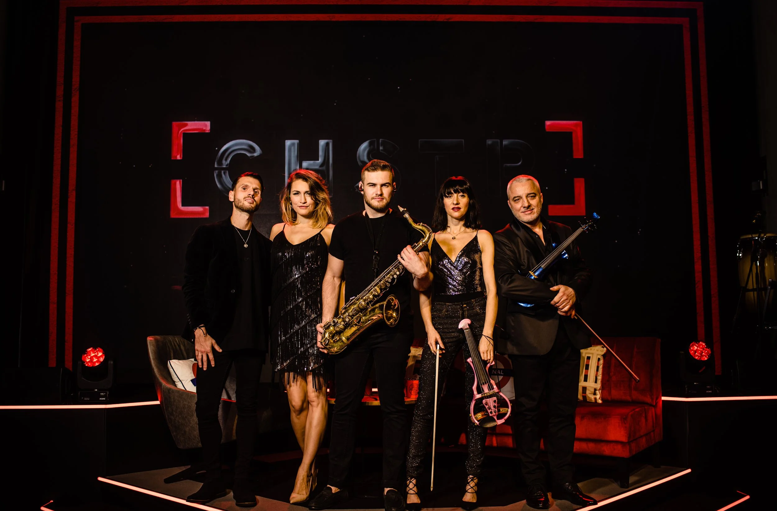 Group of five musicians on stage with instruments, standing in front of a black backdrop with red neon framing and the word 'CAMEO' displayed.