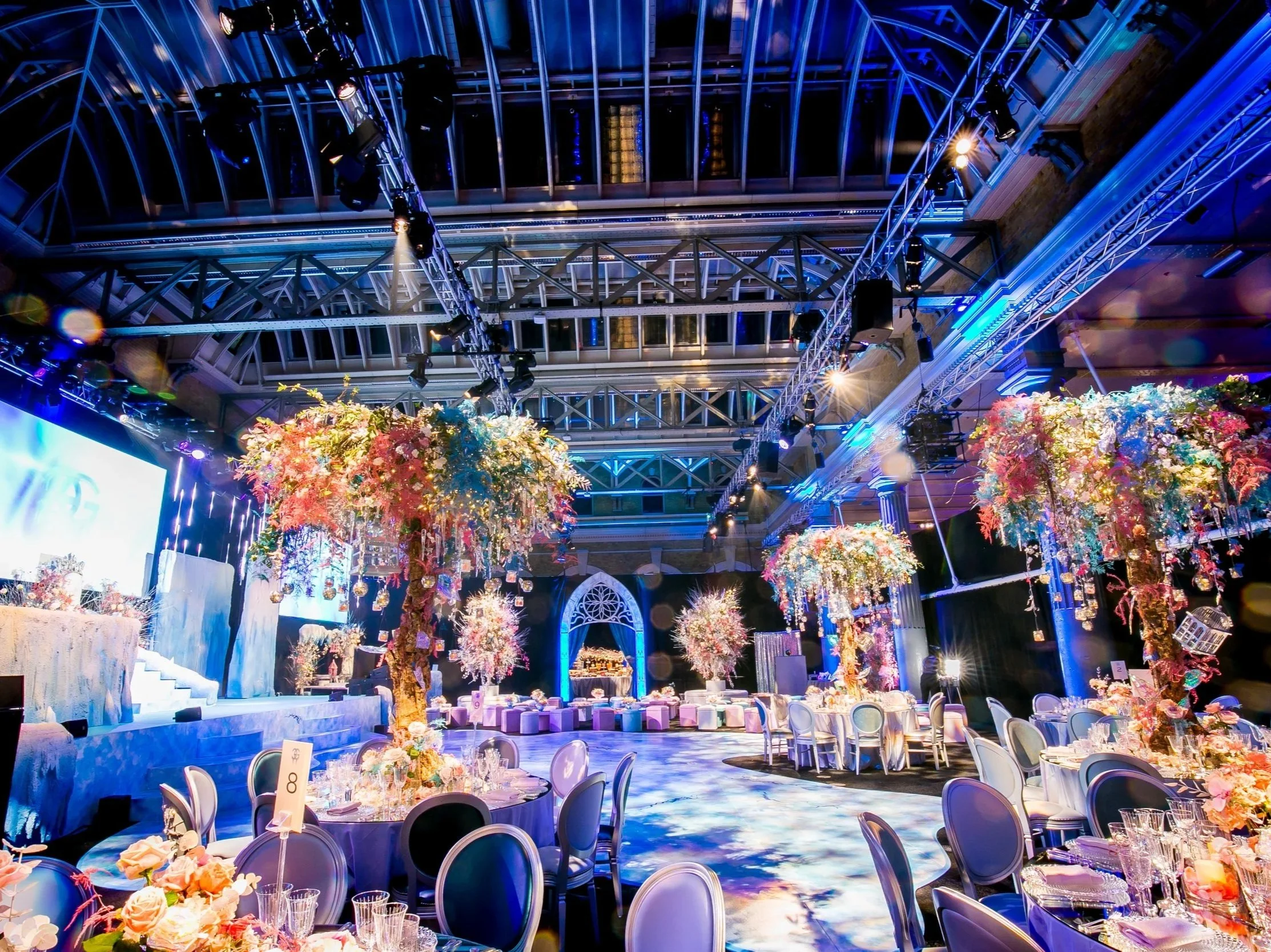 A large, elegant indoor event space decorated with colorful floral arrangements, illuminated with blue and purple lighting, and set up with round tables and chairs for a celebration or banquet. ICE PALACE BAT MITZVAH at Old Billingsgate 