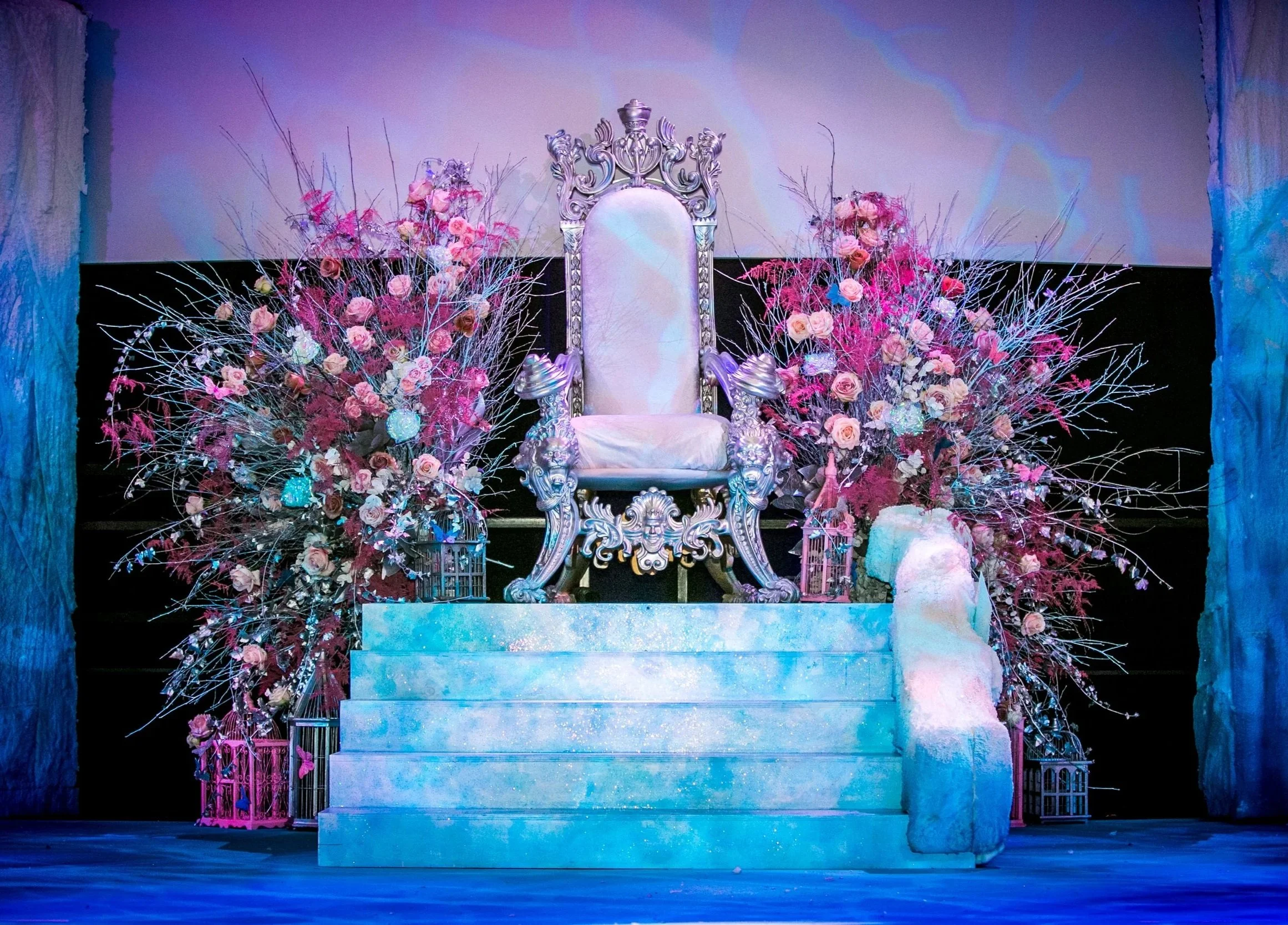 A decorative throne with ornate carvings, surrounded by pink and purple flowers, placed on a stage with stone steps. ICE PALACE BAT MITZVAH at Old Billingsgate 
