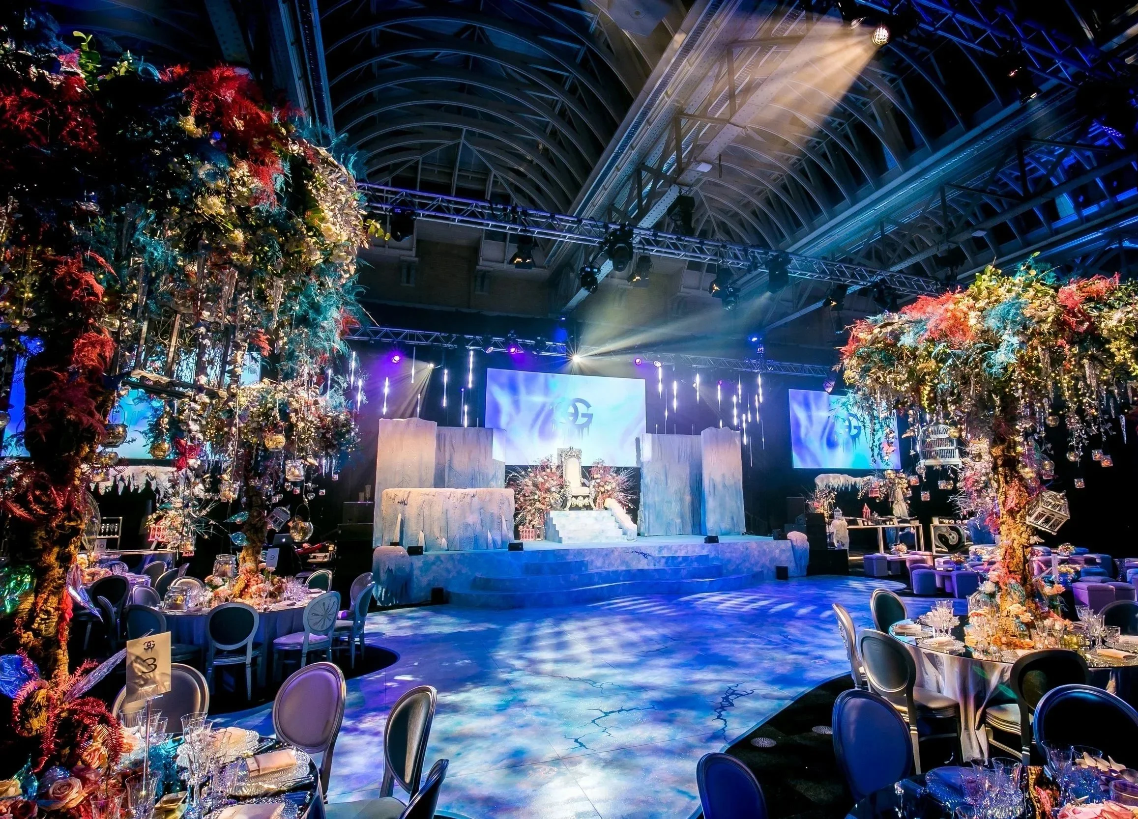 Elegant event venue decorated with colorful floral arrangements and draped lighting, featuring a stage with a large screen and seating area. ICE PALACE BAT MITZVAH at Old Billingsgate 