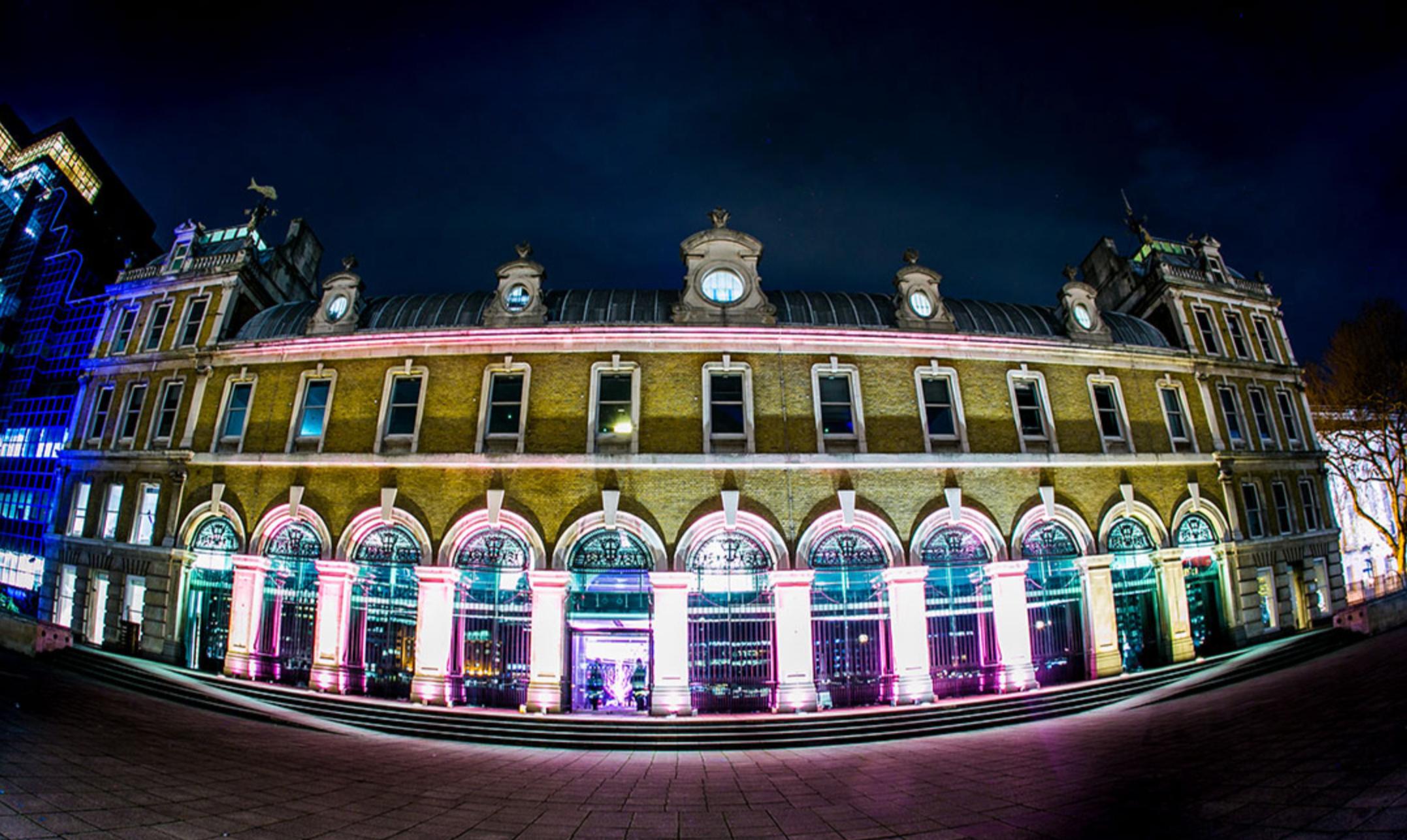 A historic building illuminated with colorful lights at night, featuring arched windows and decorative roof details, with modern buildings in the background. ICE PALACE BAT MITZVAH at Old Billingsgate 
