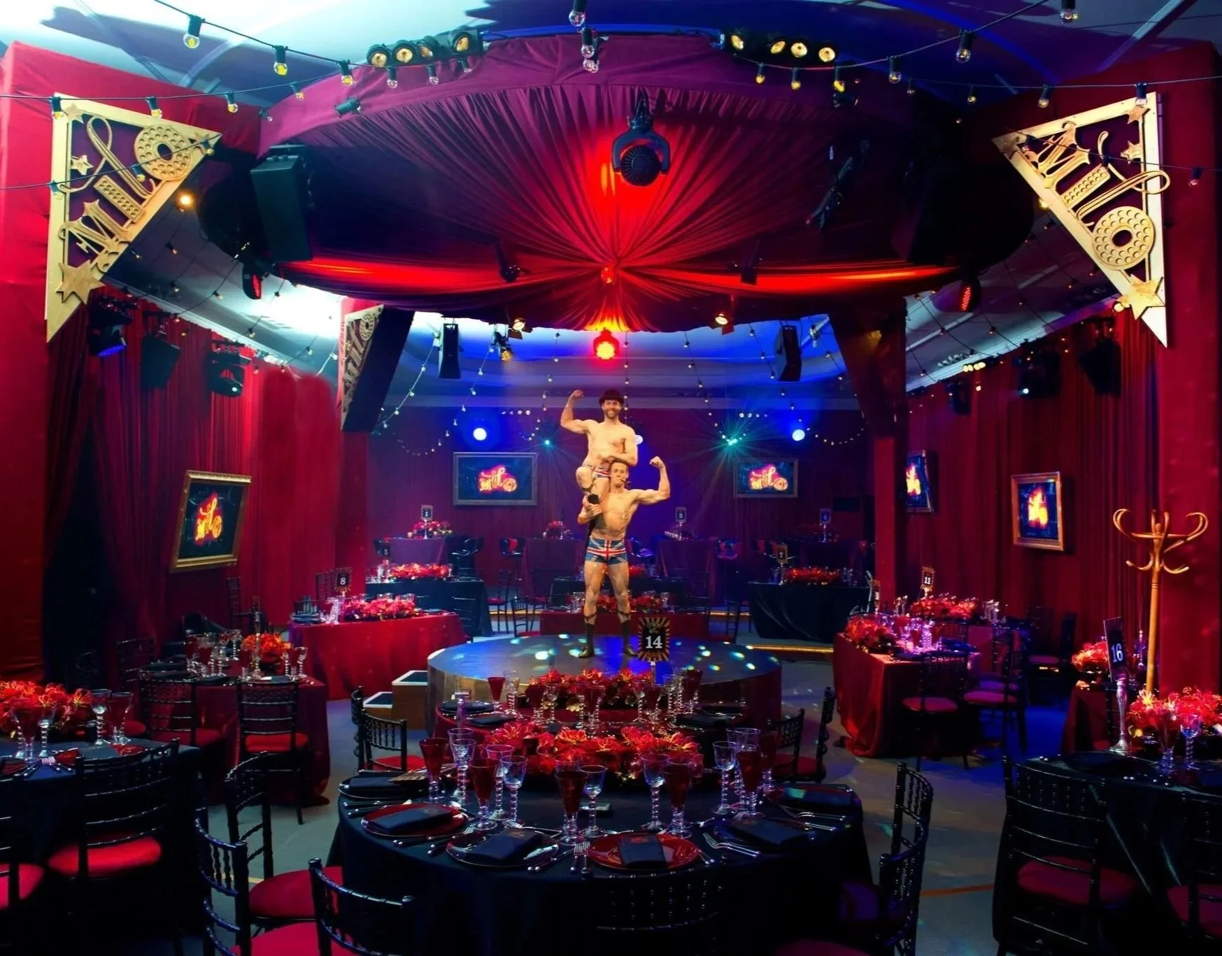 Circular stage with two male performers, one standing and the other crouching, wearing patriotic-themed outfits, in a decorated event space with red curtains, tables set with red and black tableware, and a star-shaped spotlight on the ceiling.
